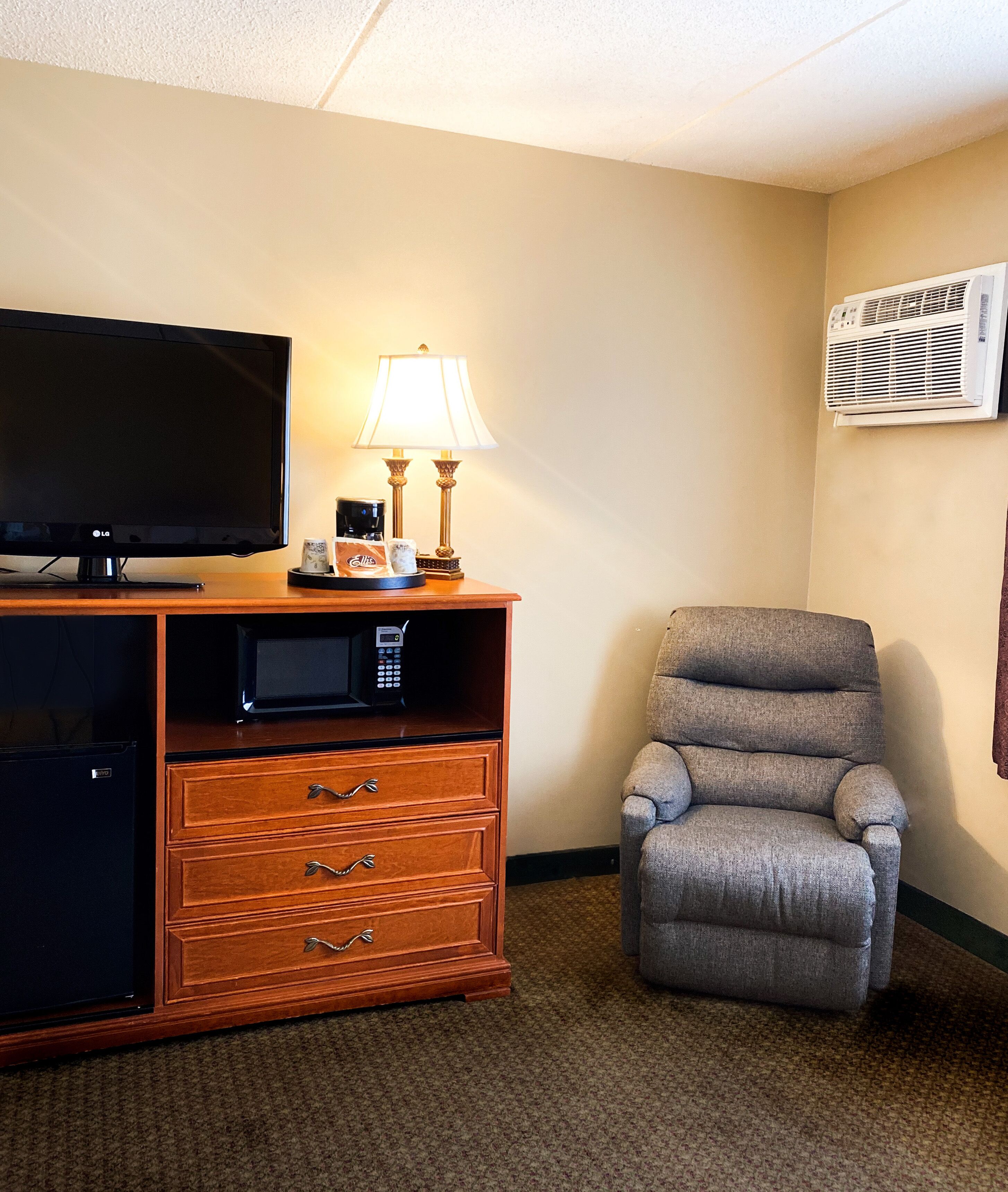 A hotel room with a flat-screen TV, a lamp, a microwave, a small refrigerator, and a grey armchair.