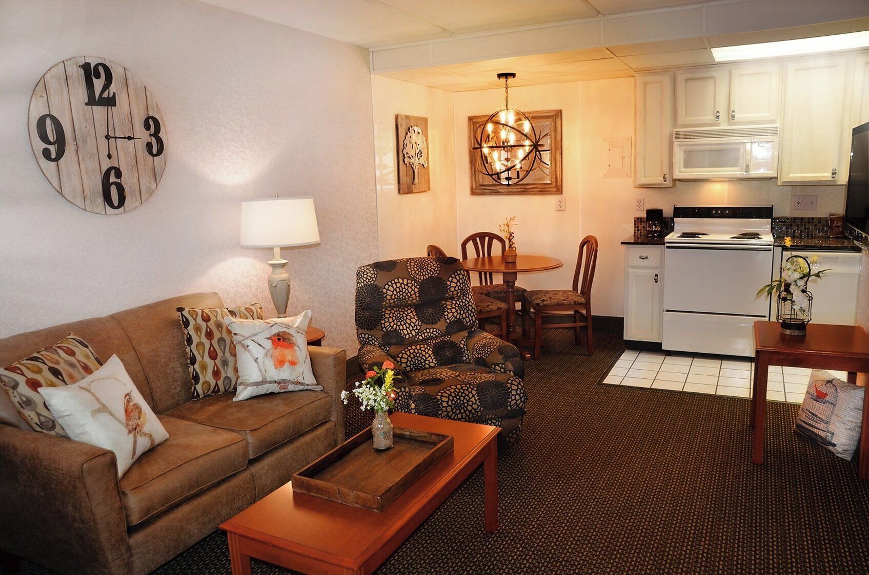 A living room with a brown sofa, patterned armchair, wooden coffee table, and a dining area with a wooden table and chairs. A kitchen with white cabinets and appliances is visible in the background.