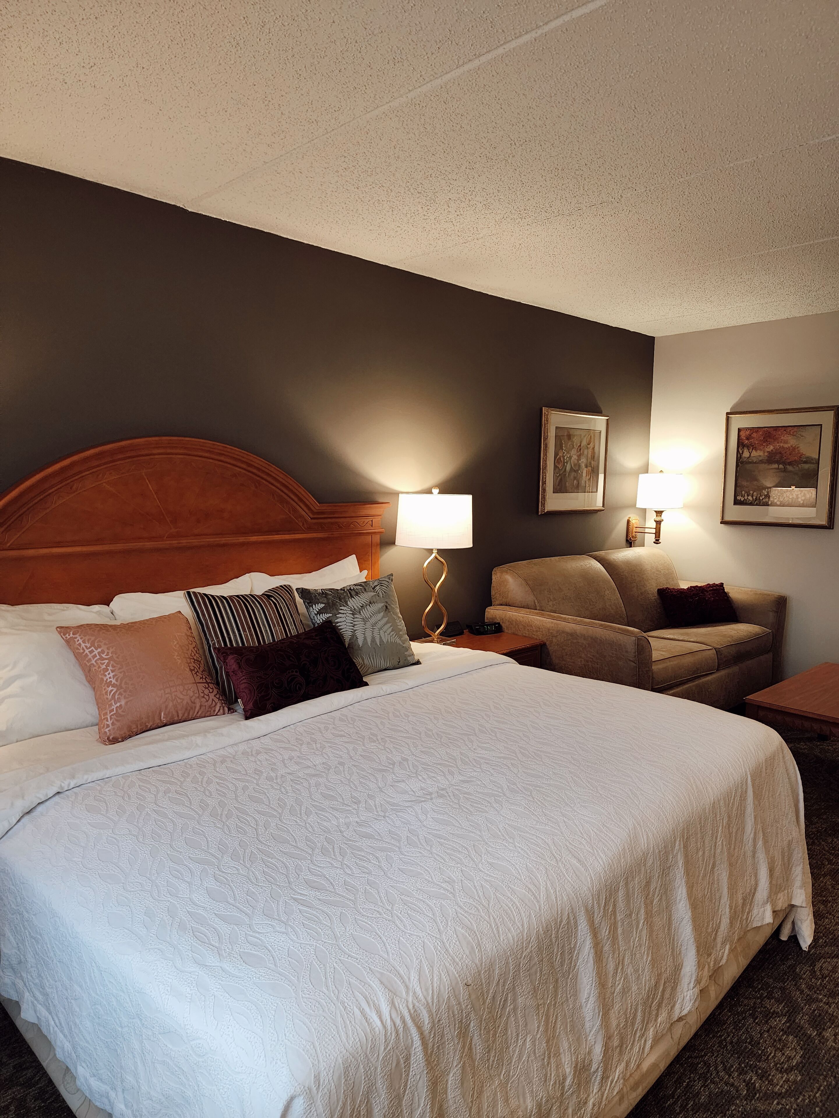 Habitación de hotel con una cama grande, cabecera de madera, dos mesitas de noche con lámparas, un sofá de cuero marrón y dos cuadros en la pared.