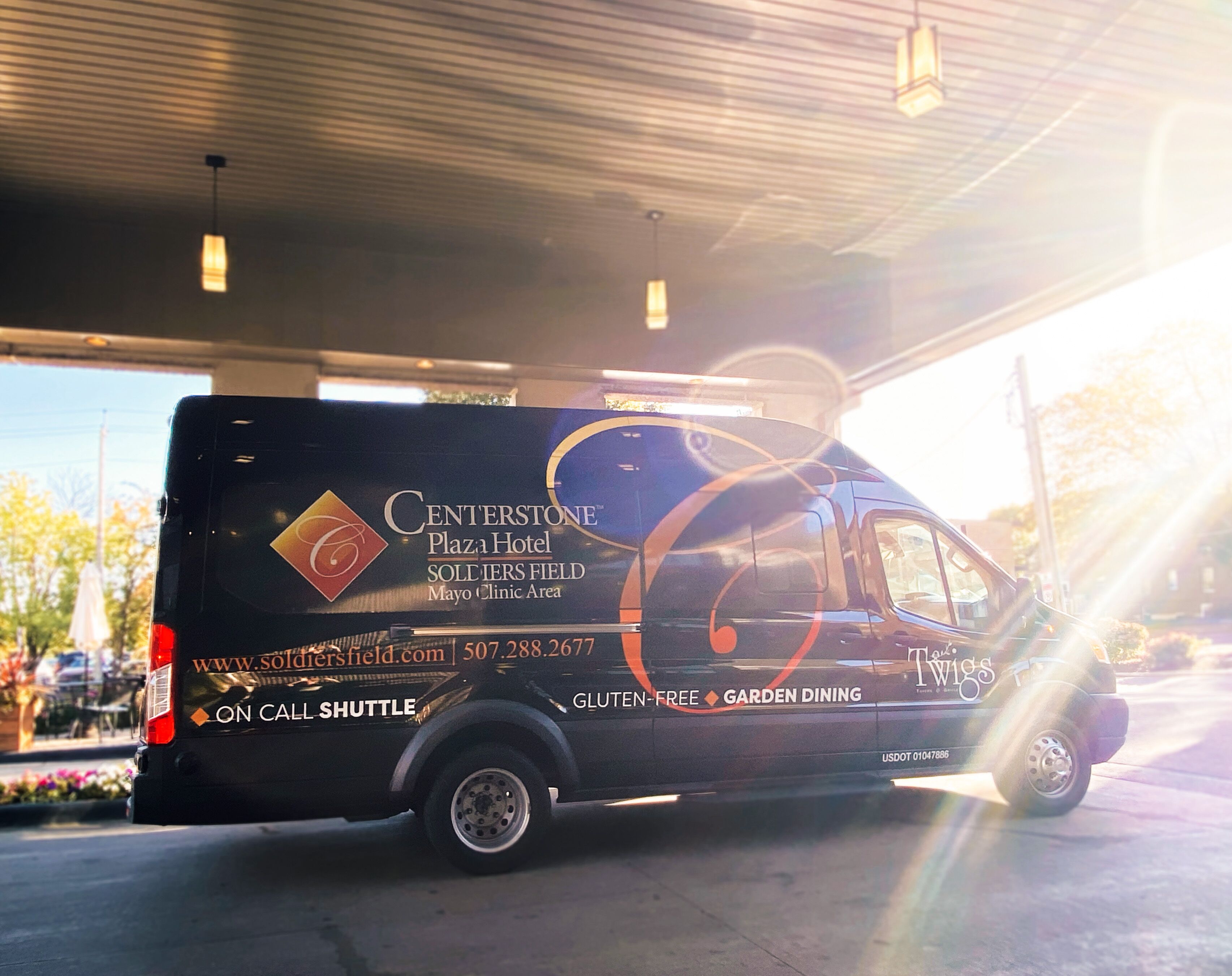 A shuttle bus parked at a gas station with a sign advertising Centerstone Plaza Hotel and services like on-call shuttle and gluten-free garden dining.