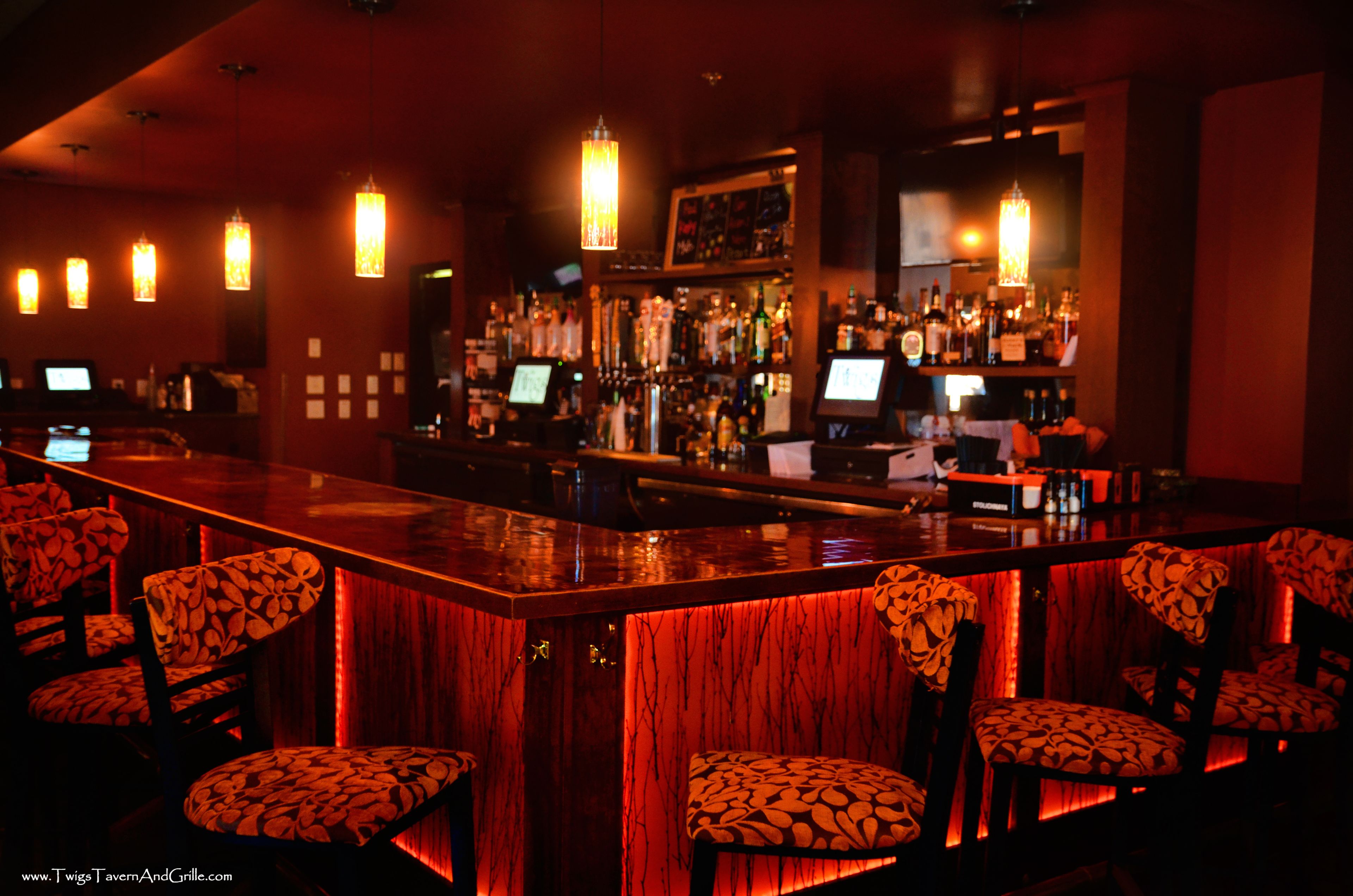 A bar with a red-lit bar counter, patterned chairs, and a well-stocked liquor shelf.
