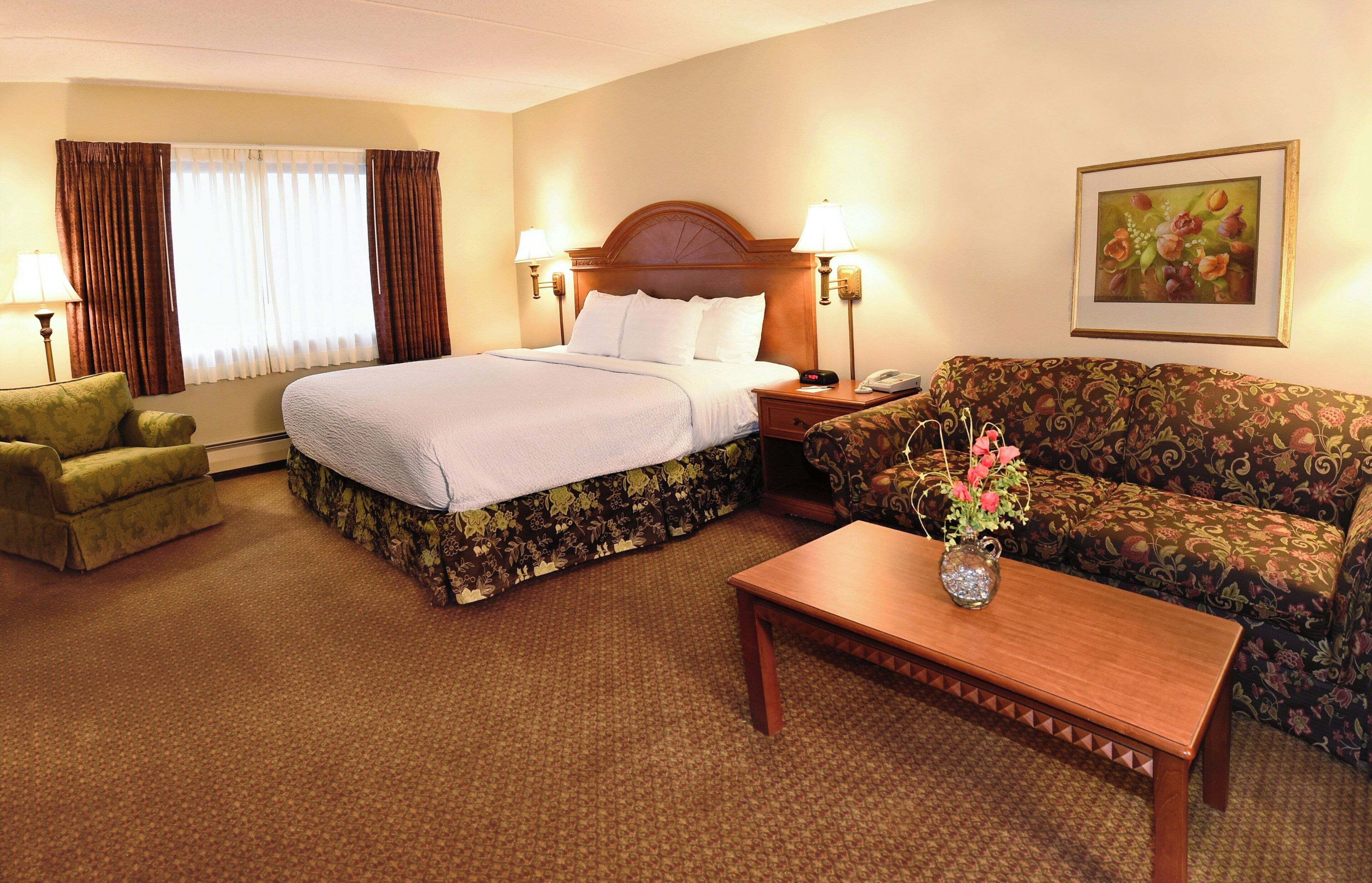 A hotel room with a bed, a sofa, a small table, a chair, and a painting on the wall.