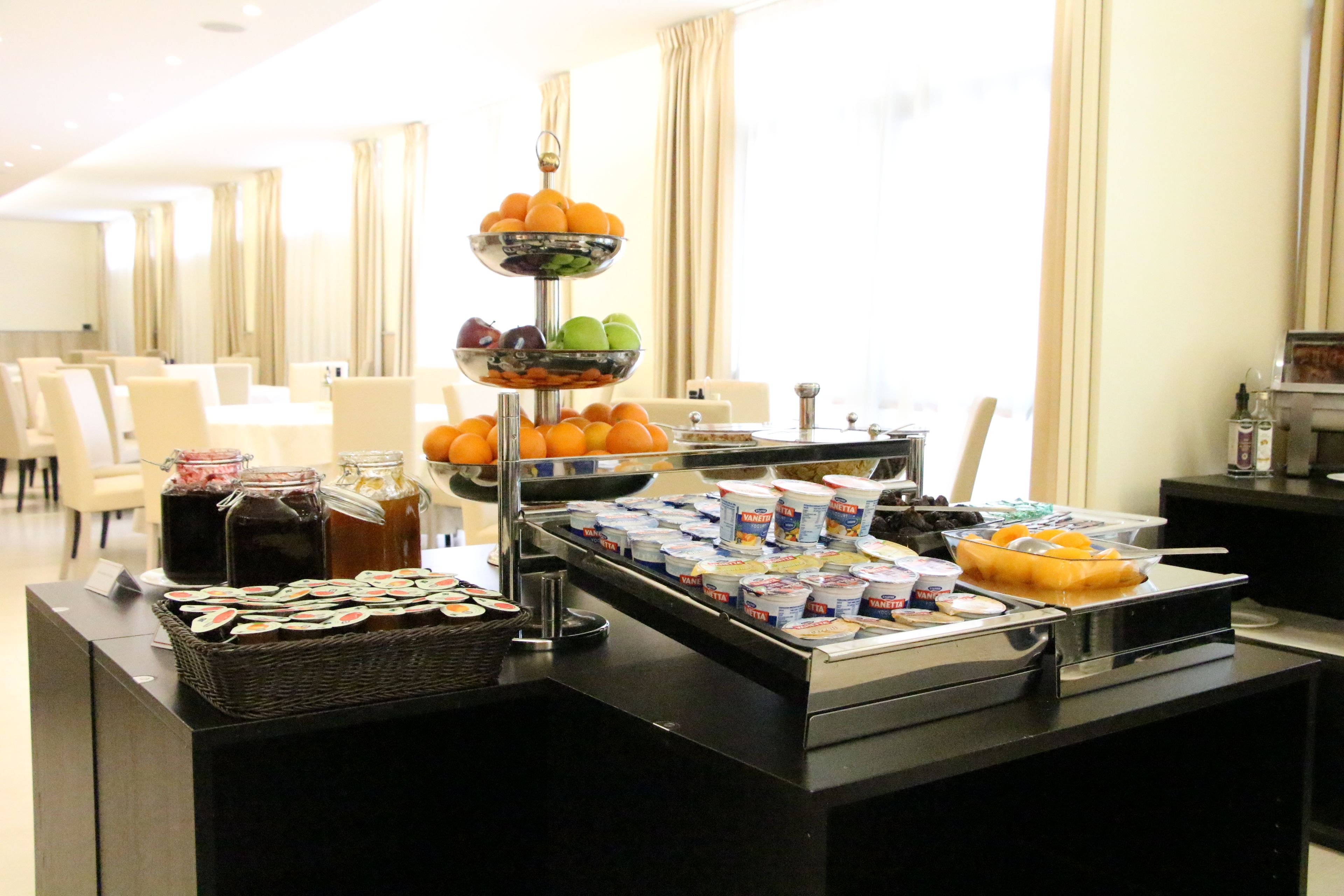 A buffet setup with various food items and beverages, including fruits, yogurt, and juices, in a well-lit room with a seating area in the background.
