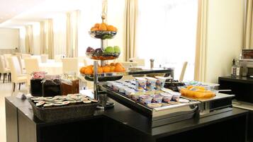 A buffet setup with various food items and beverages, including fruits, yogurt, and juices, in a well-lit room with a seating area in the background.