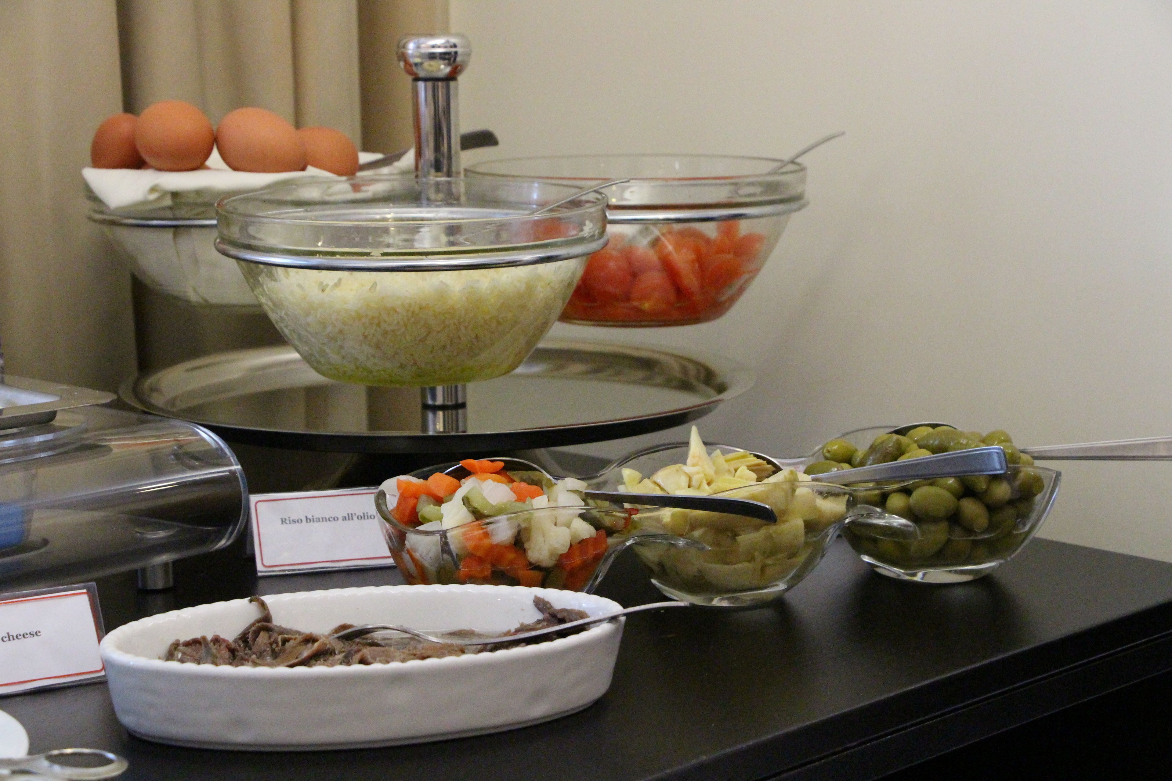 A buffet with various food options including a bowl of shredded cheese, a bowl of sliced vegetables, and a bowl of olives.