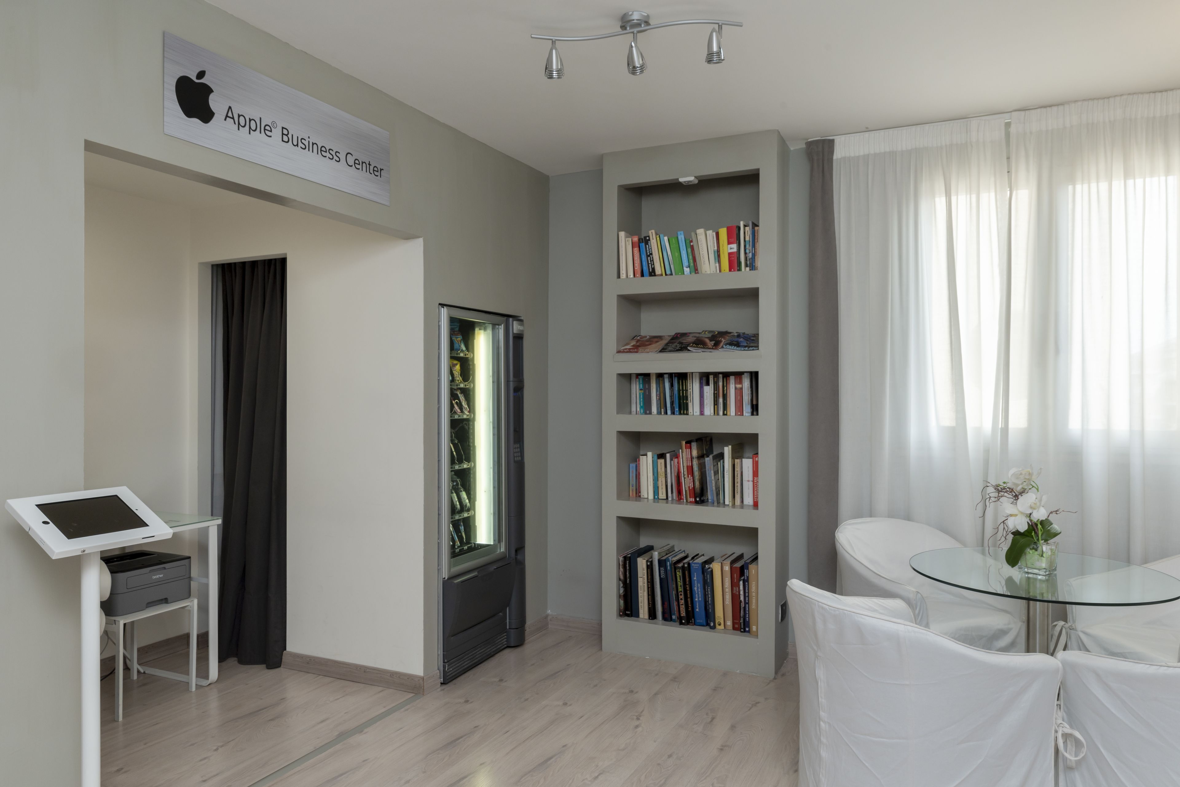 A modern Apple Business Center with a flat-screen TV, a bookshelf, and a glass table with chairs.