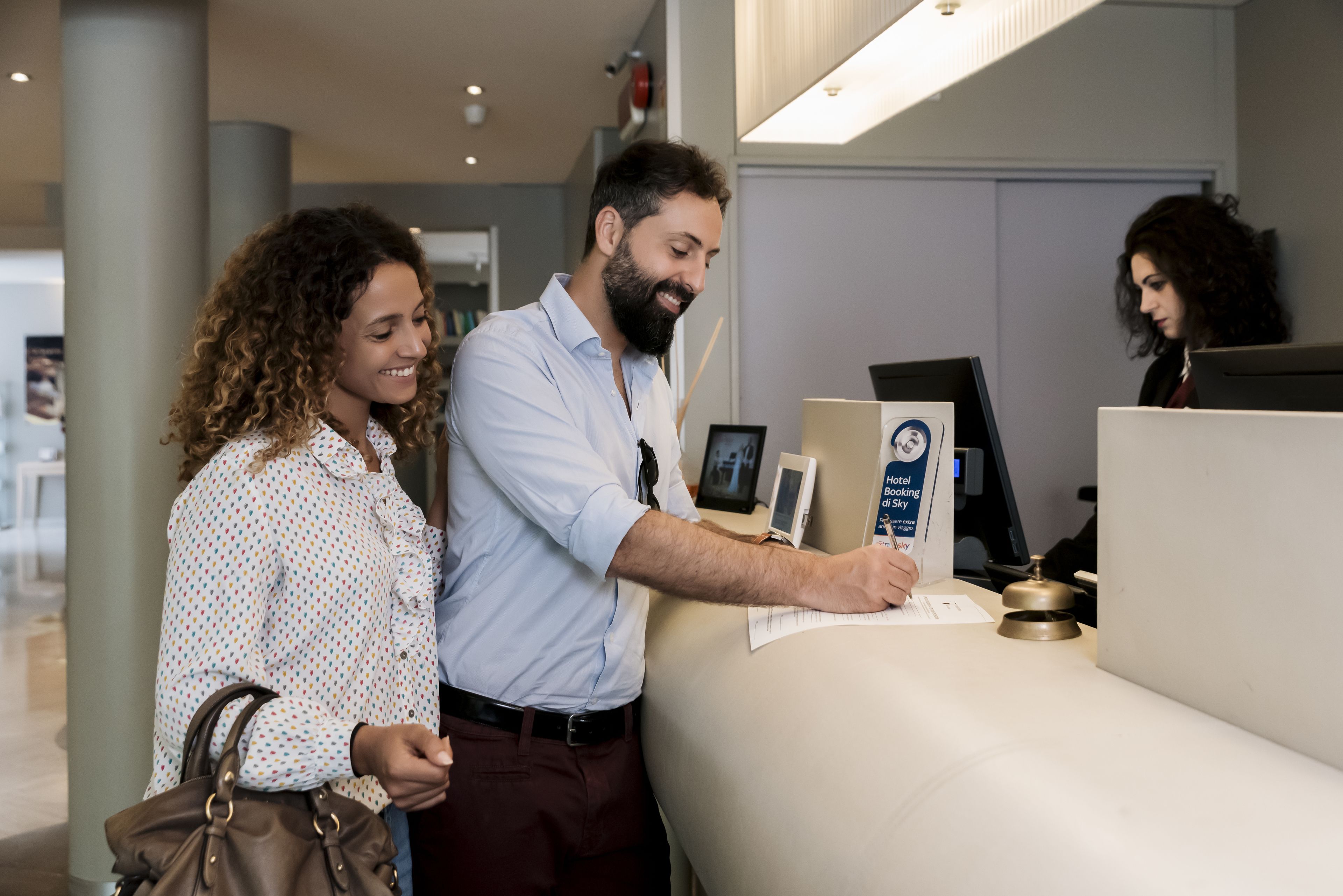 A reception desk with a man and a woman standing nearby, a sign indicating hotel amenities, and a computer monitor.