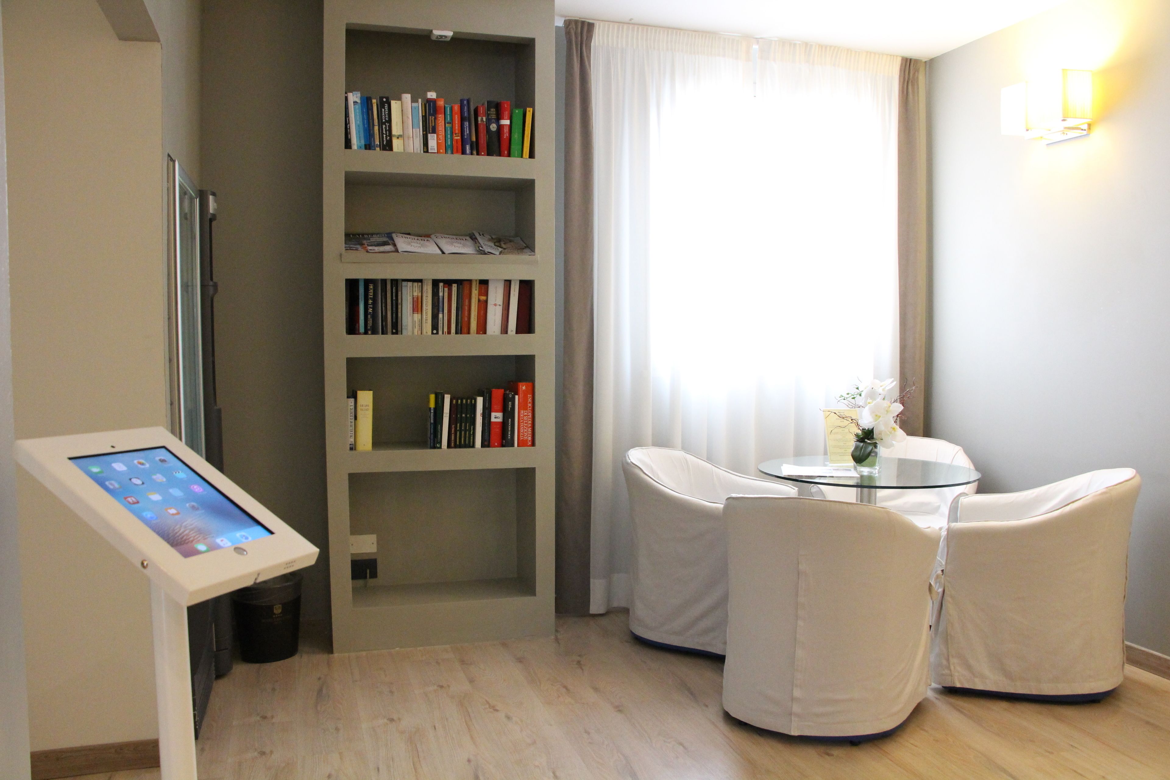 Quarto de hotel com um quiosque digital, uma estante de livros, uma mesa redonda com duas cadeiras e uma janela com cortinas.