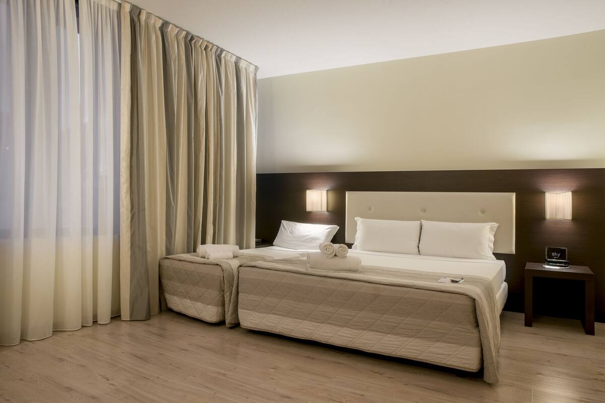 Habitación de hotel con una cama grande, dos mesitas de noche, una lámpara y una ventana con cortinas.