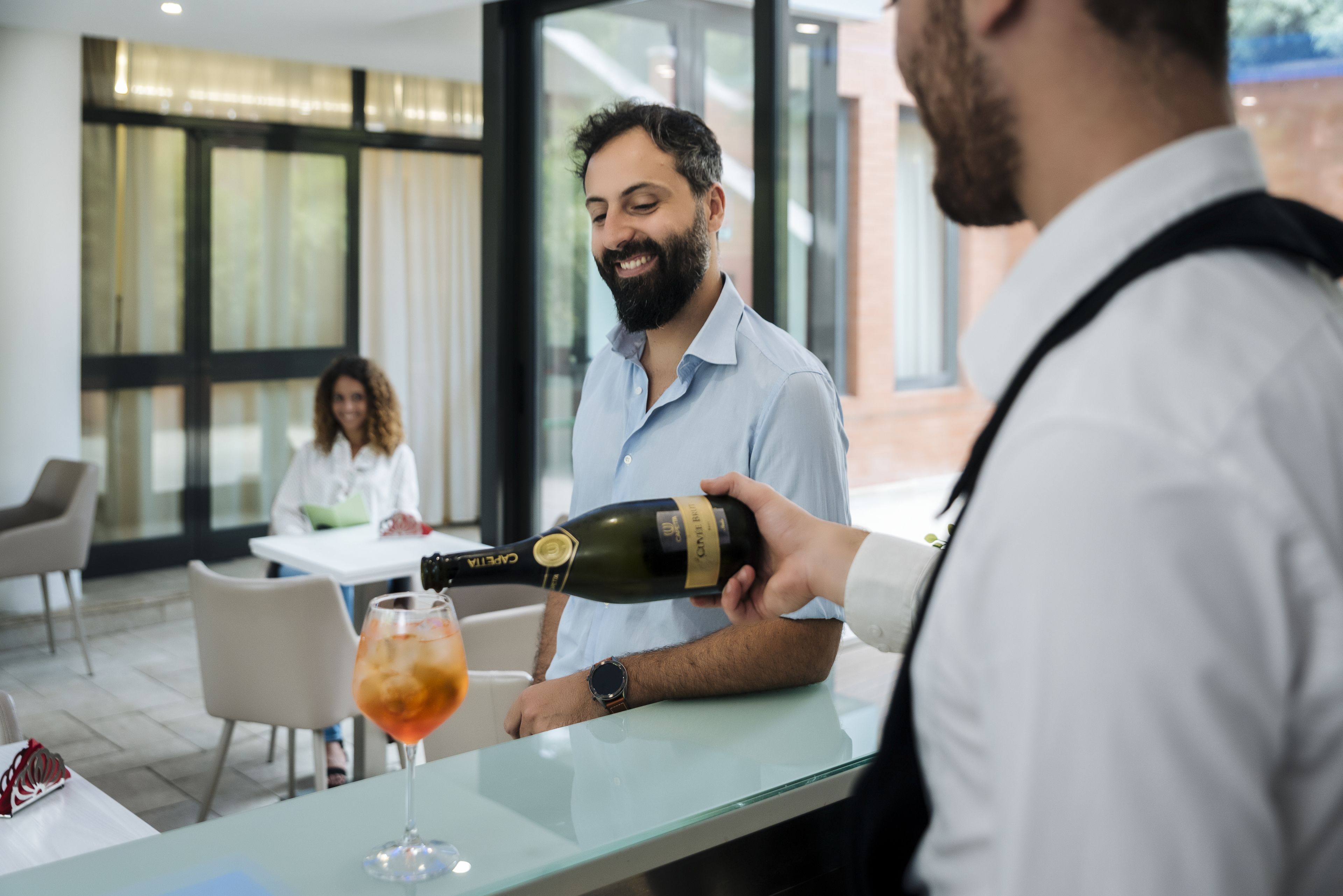 En man som häller upp en drink från en flaska i en bar, med ett glas vin på bardisken.