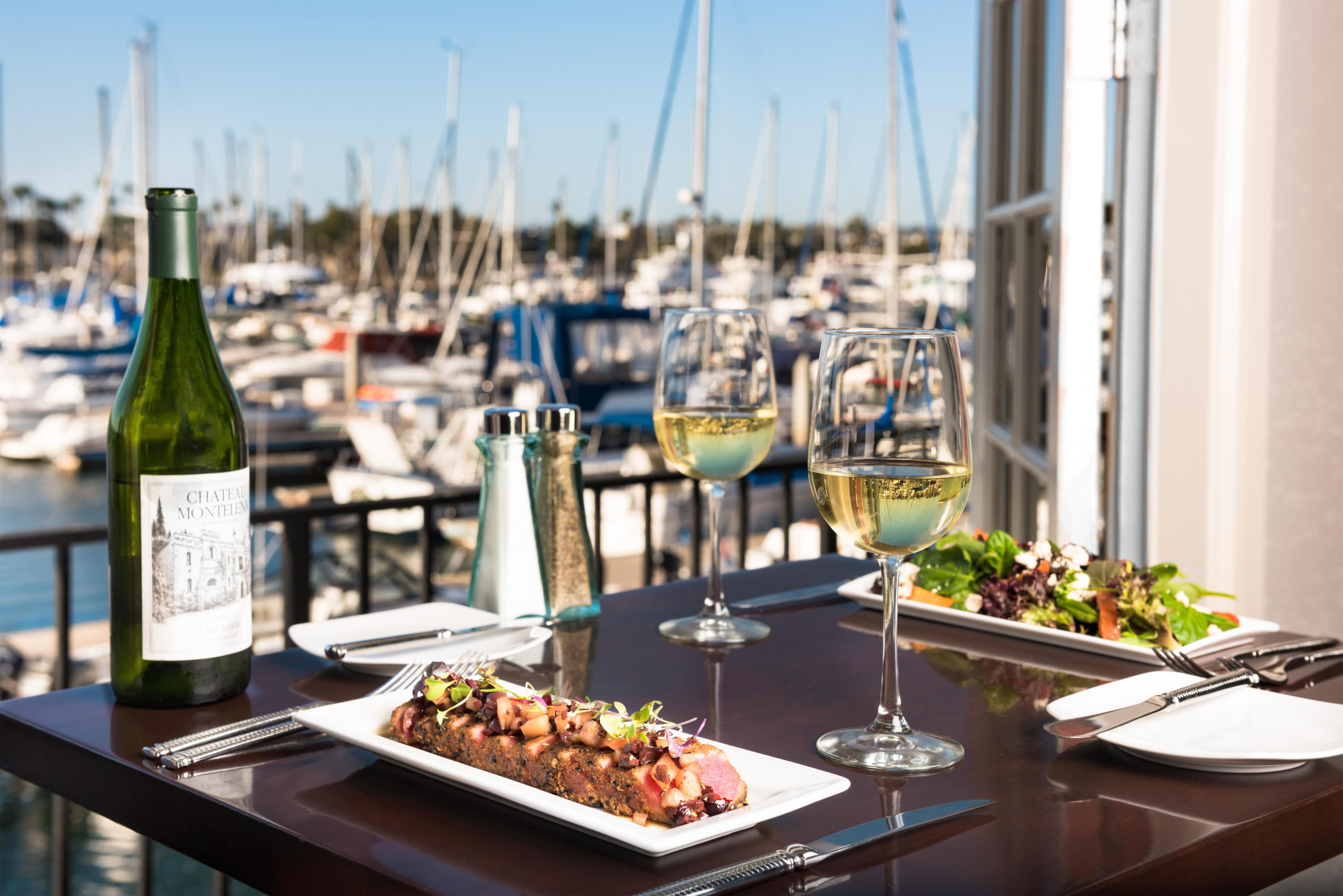 A dining table with a bottle of wine, two glasses, a plate of food, and a view of a marina with boats.