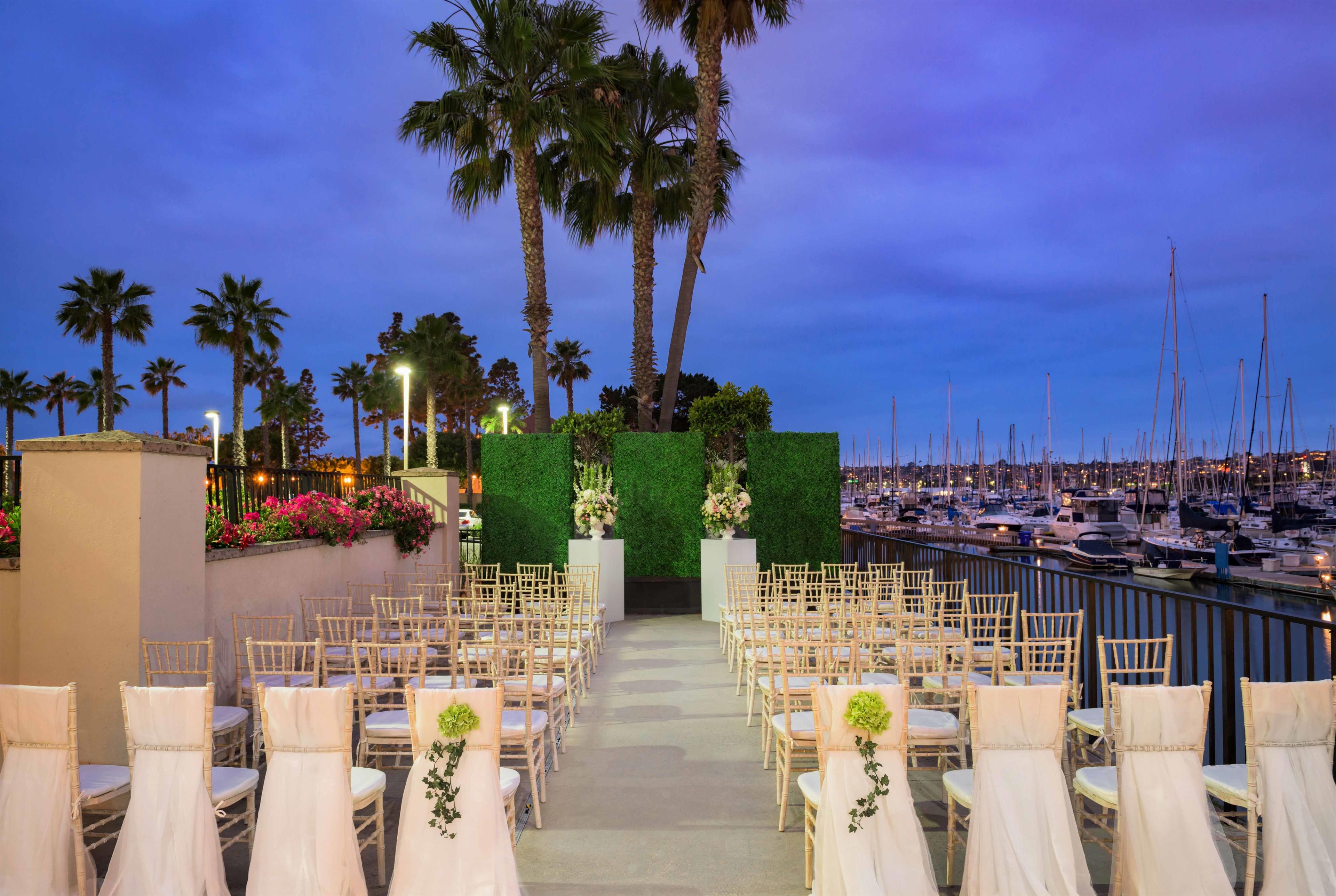 Un'area salotto all'aperto con file di sedie e tavoli, un siepe verde e uno sfondo con un porto turistico e barche.