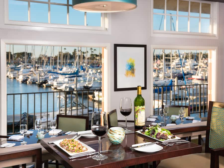 A dining table set with plates, glasses, and a bottle of wine, overlooking a marina with boats.