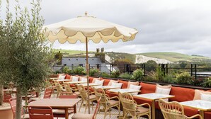 An outdoor seating area with tables and chairs, a large umbrella, and a view of the countryside. - The White Hart - A Heartwood Inn (Lewes)