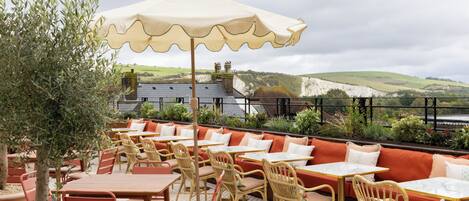 An outdoor seating area with tables and chairs, a large umbrella, and a view of the countryside.