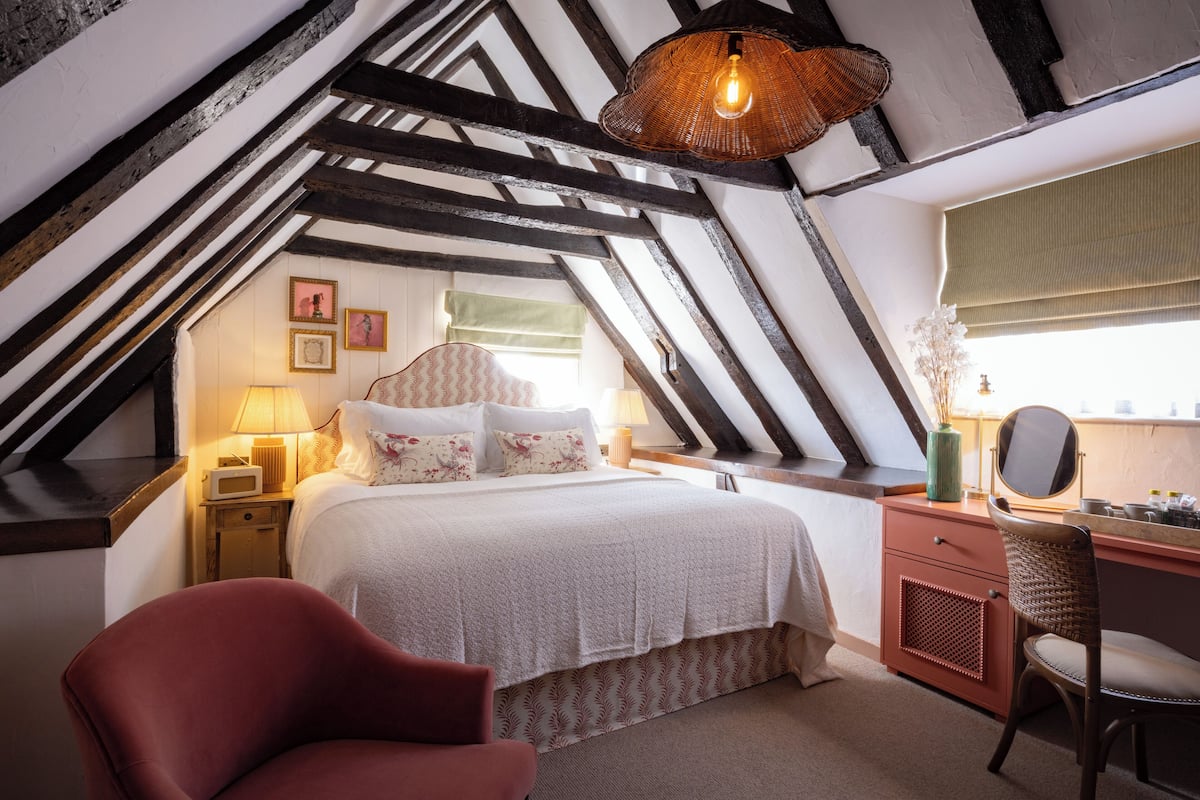 Une chambre avec un plafond en pente, un lit recouvert d’une literie à motifs floraux, un fauteuil rouge, un bureau en bois avec un miroir et un luminaire suspendu.