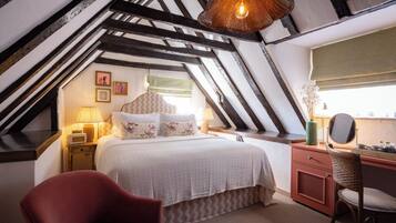 A bedroom with a sloped ceiling, a bed with floral bedding, a red armchair, a wooden desk with a mirror, and a hanging light fixture.