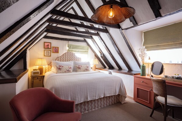Une chambre avec un plafond en pente, un lit recouvert d’une literie à motifs floraux, un fauteuil rouge, un bureau en bois avec un miroir et un luminaire suspendu.