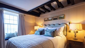 A neatly made bed with a quilted cover, a bedside table with a lamp, and a window with curtains. - The White Hart - A Heartwood Inn (Lewes)