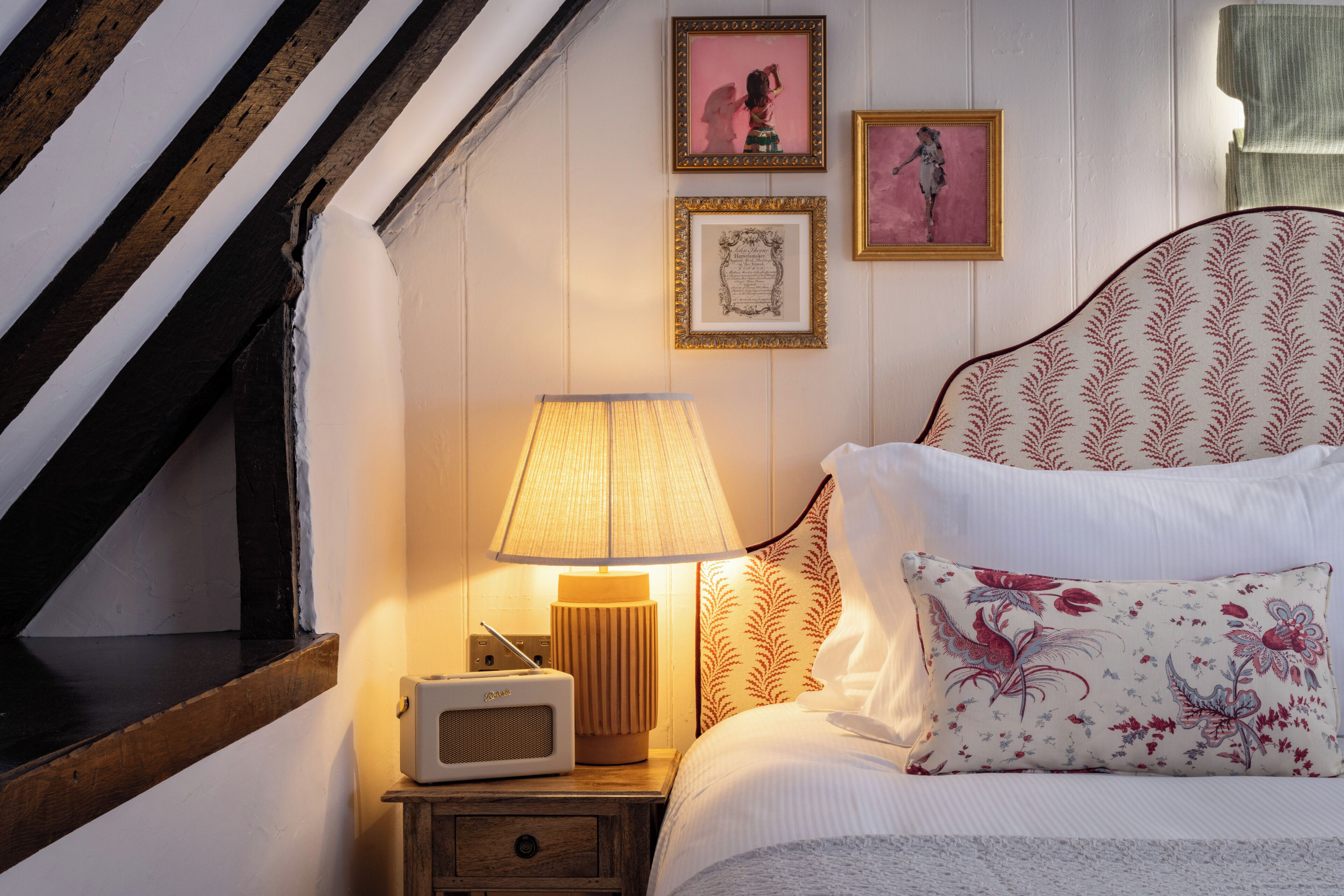 Een slaapkamer met een houten plafond, een nachtkastje met een lamp en radio, een bed met bloemenkussens en ingelijste foto's aan de muur.