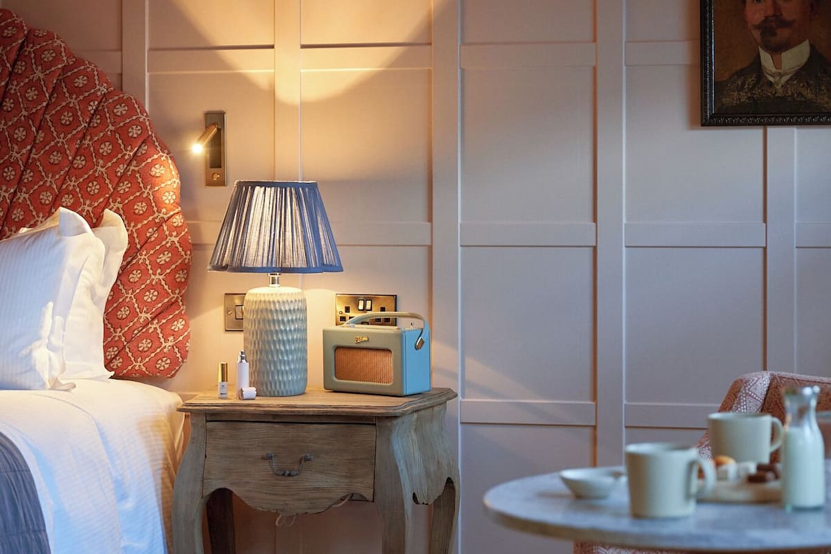 Une chambre d’hôtel comprenant un lit, une table de chevet avec une lampe, une radio, une petite table avec des tasses et un pichet, et une chaise.