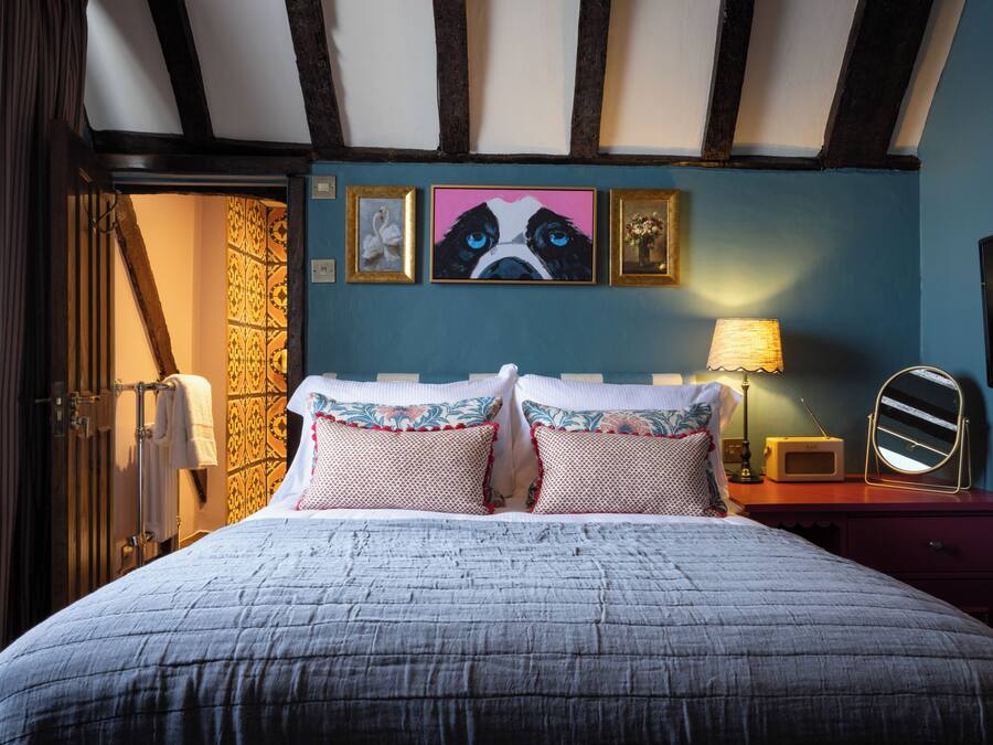 A bedroom with a large bed, a wooden ceiling, a wall with framed artwork, a bedside table with a lamp, and a mirror.