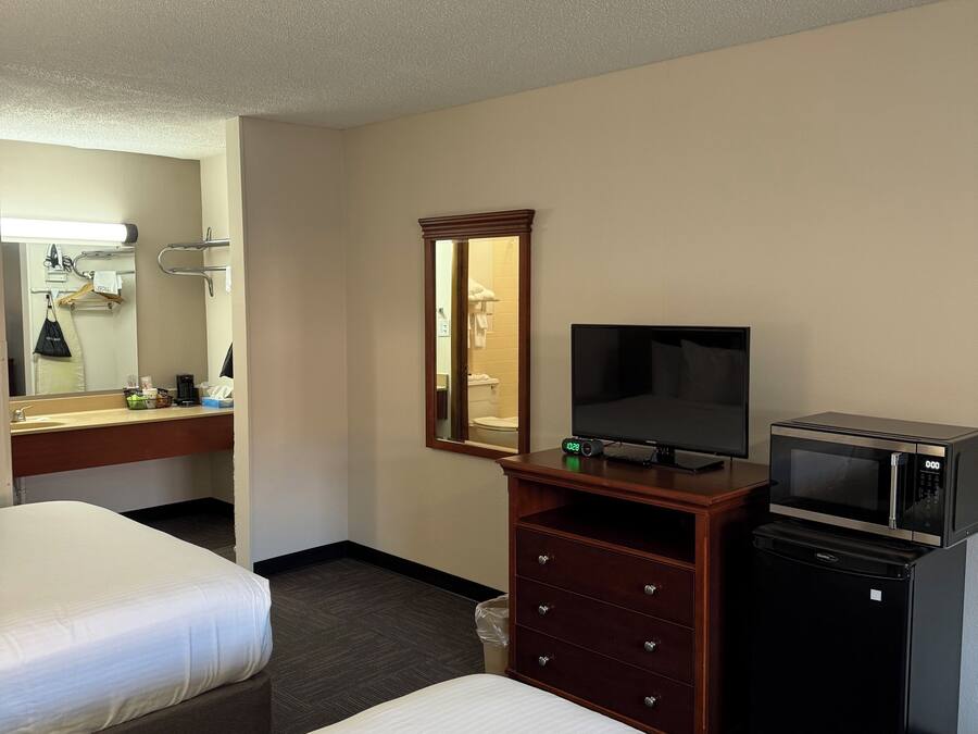 A hotel room with two beds, a TV, a microwave, a small refrigerator, a wooden dresser, a mirror, and a bathroom visible through an open door.