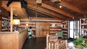 A rustic wooden interior with a reception desk, wooden chairs, a large plant, and a refrigerator.