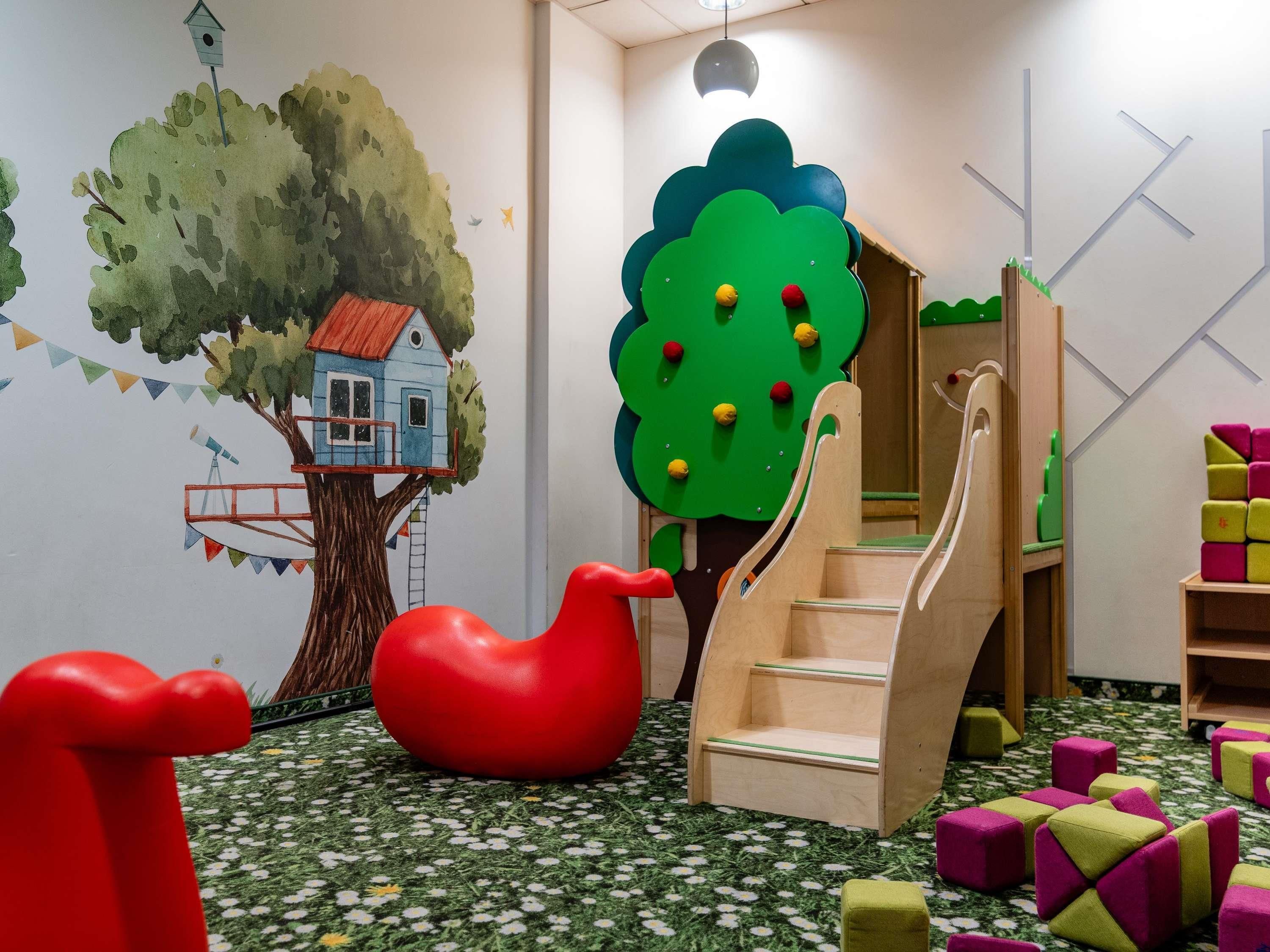 Une salle de jeux pour enfants avec une fresque représentant une cabane dans les arbres, un toboggan et des blocs de construction colorés.