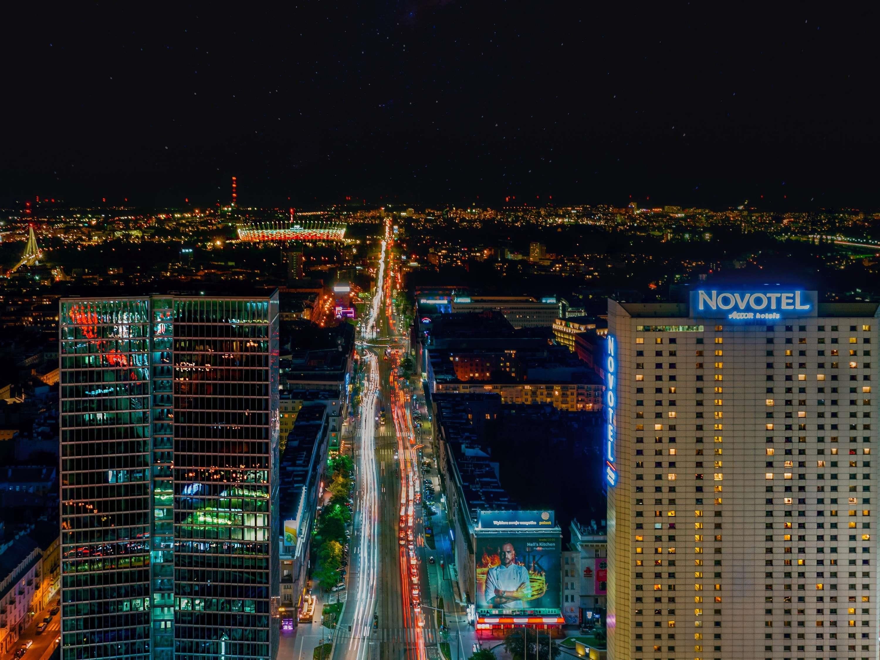 Un hotel de gran altura con un llamativo letrero 'NOVOTEL', una calle concurrida con estelas de luz y un paisaje urbano al fondo.