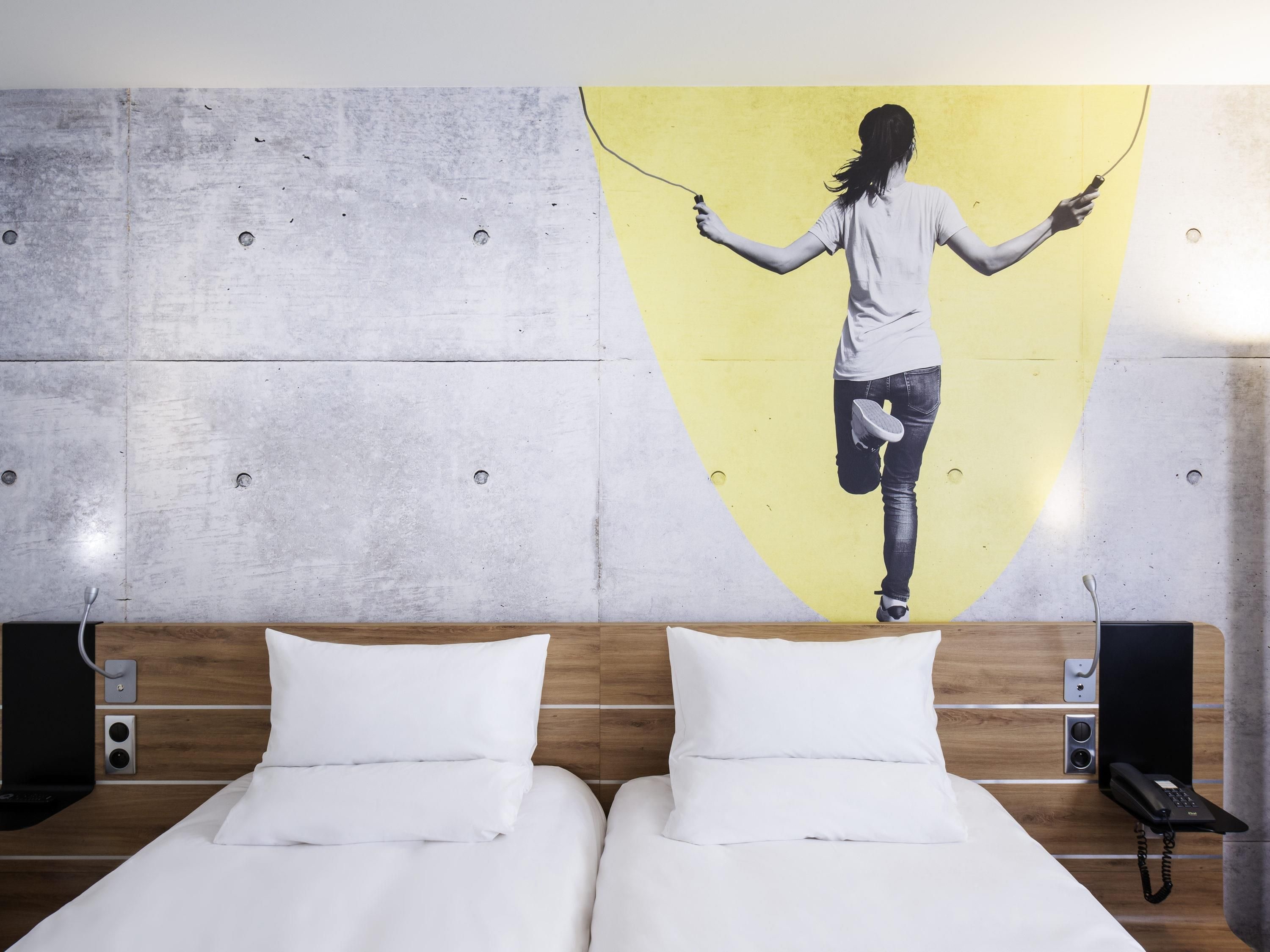A hotel room with a bed, two pillows, a wooden headboard, a wall-mounted phone, and a large painting of a person jumping rope.
