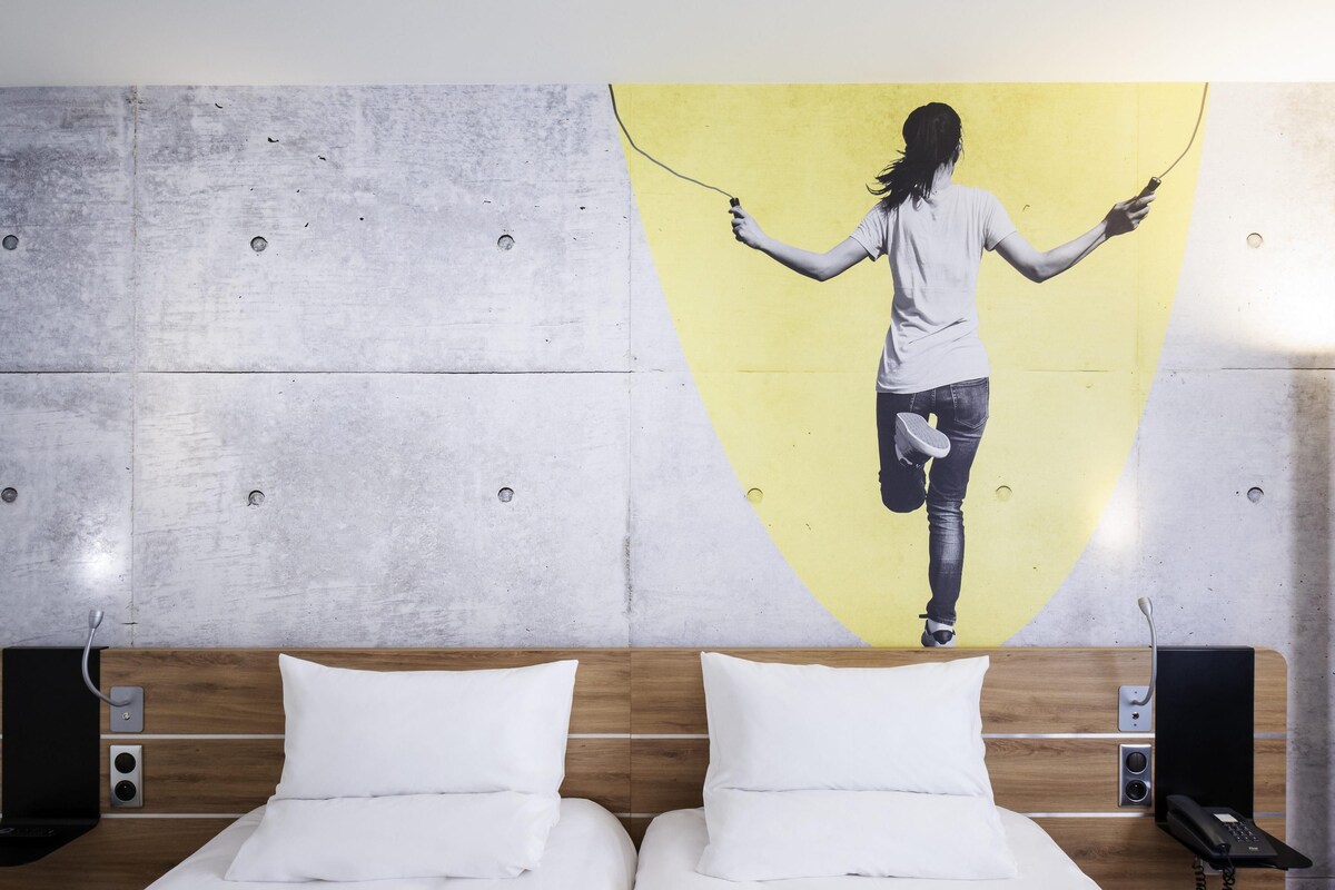 A hotel room with a bed, two pillows, a wooden headboard, a wall-mounted phone, and a large painting of a person jumping rope.