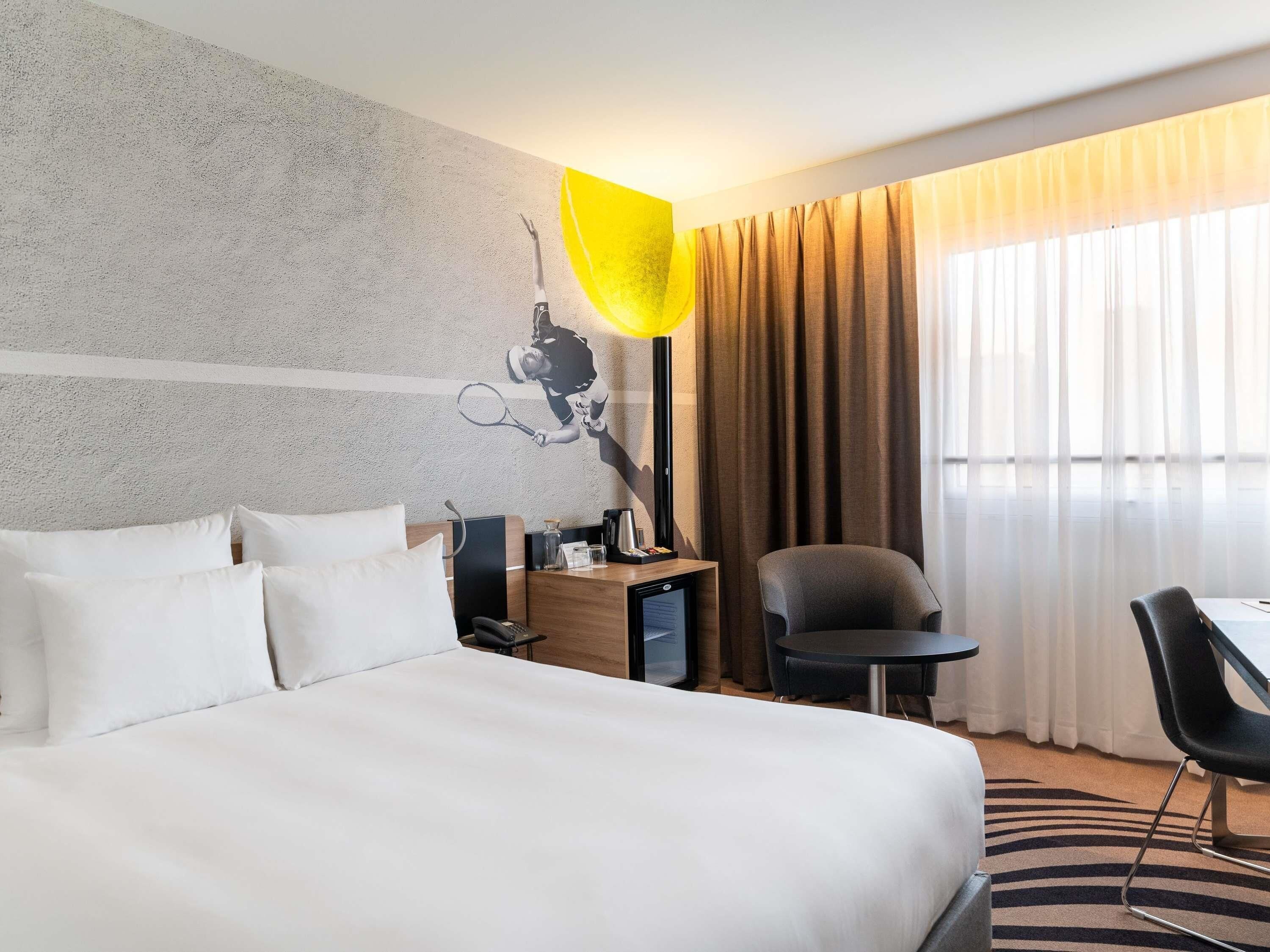A hotel room with a large bed, a desk with a chair, a lamp, and a view of the outdoors through sheer curtains.