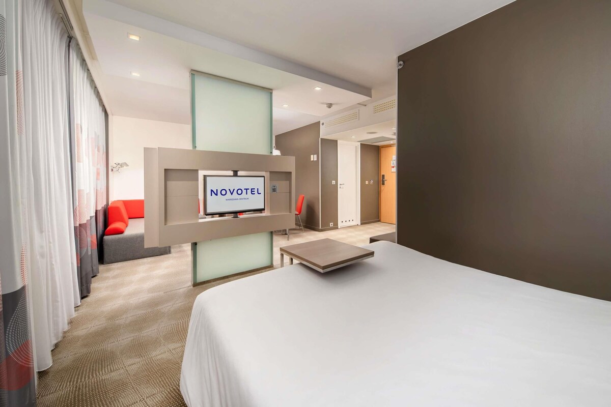 A hotel room with a bed, a TV mounted on a wall, a desk, and a door leading to another room.