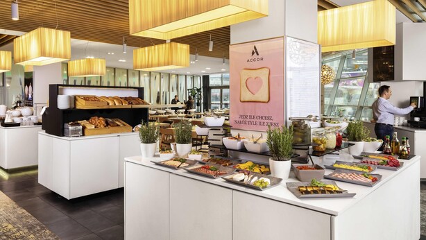 A modern breakfast area with a central counter displaying various food items, surrounded by seating and a large window. - Novotel Warszawa Centrum (Warsaw)