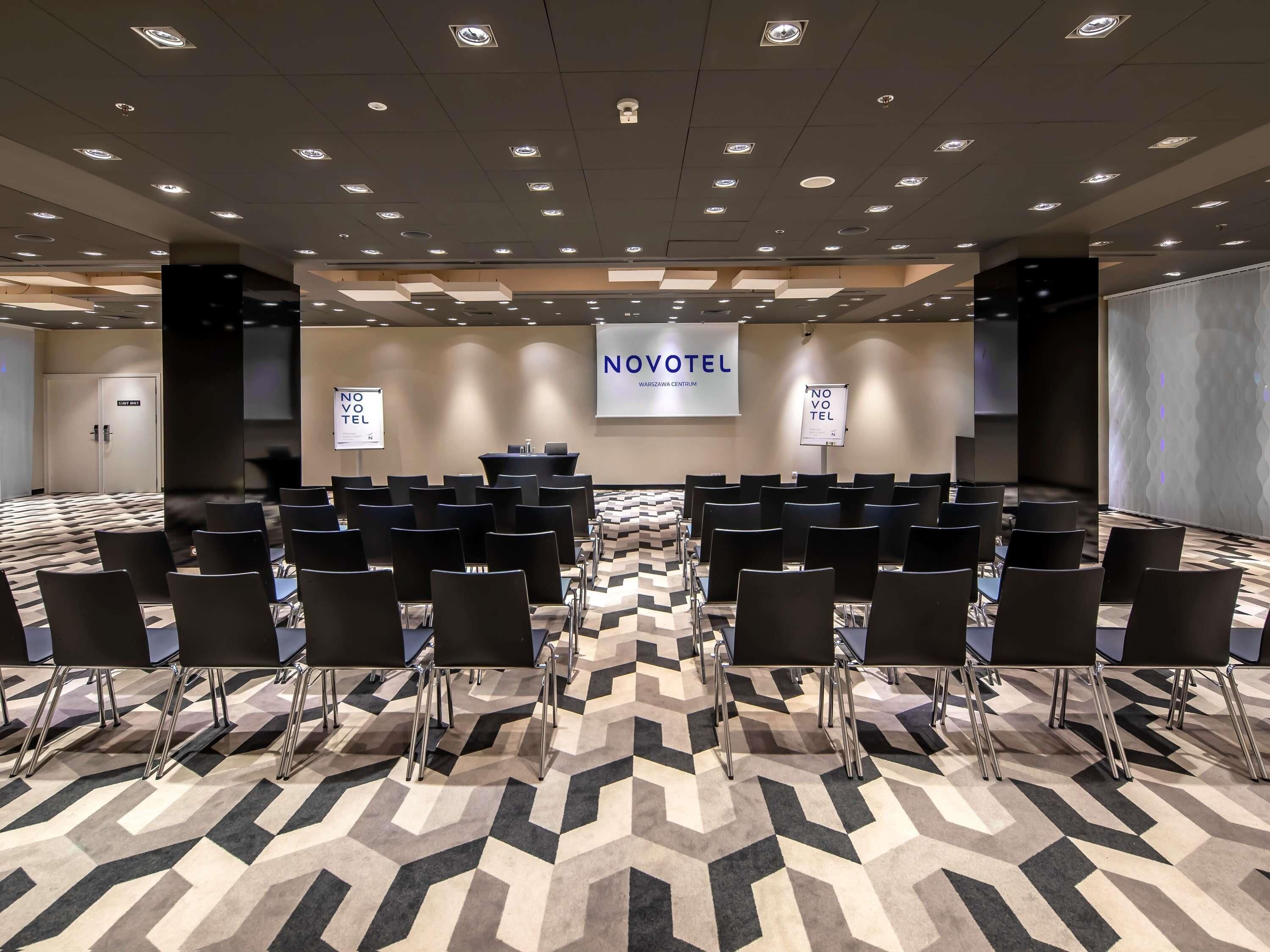 Uma sala de conferência com fileiras de cadeiras, um púlpito e uma tela exibindo 'NOVOTEL' na parede.