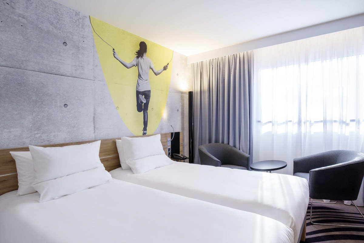 Ein Hotelzimmer mit zwei Betten, einem Bild an der Wand, einem Sessel, einem kleinen Tisch und Blick auf die Stadt durch große Fenster.
