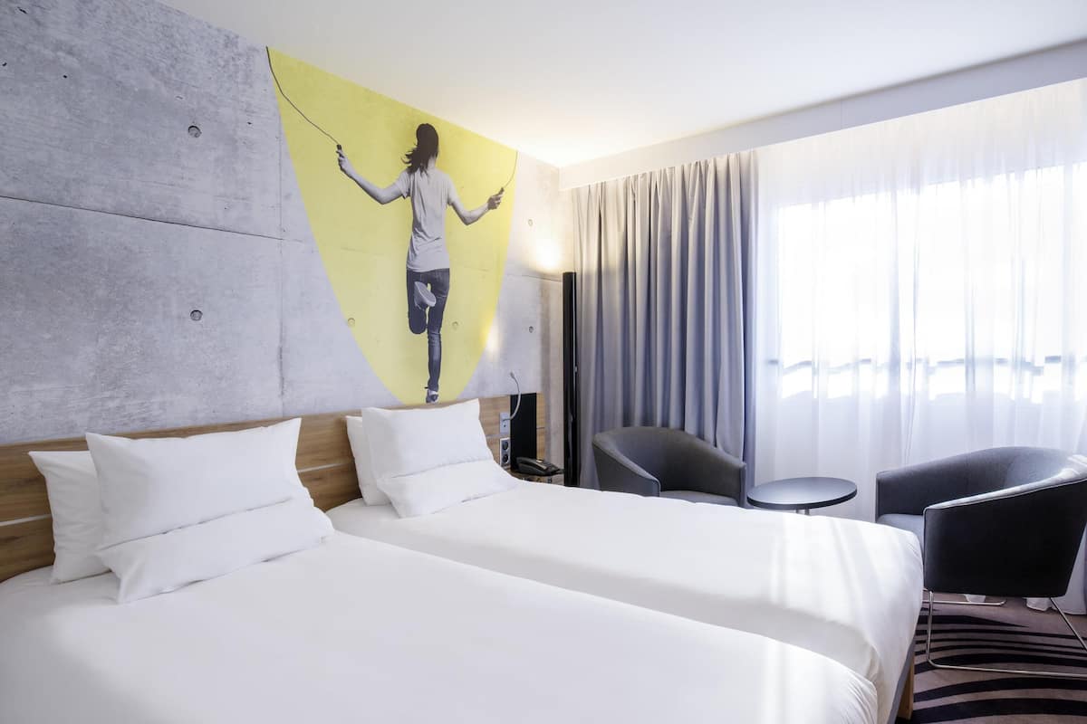 A hotel room with two beds, a painting on the wall, a chair, a small table, and a view of the city through large windows.