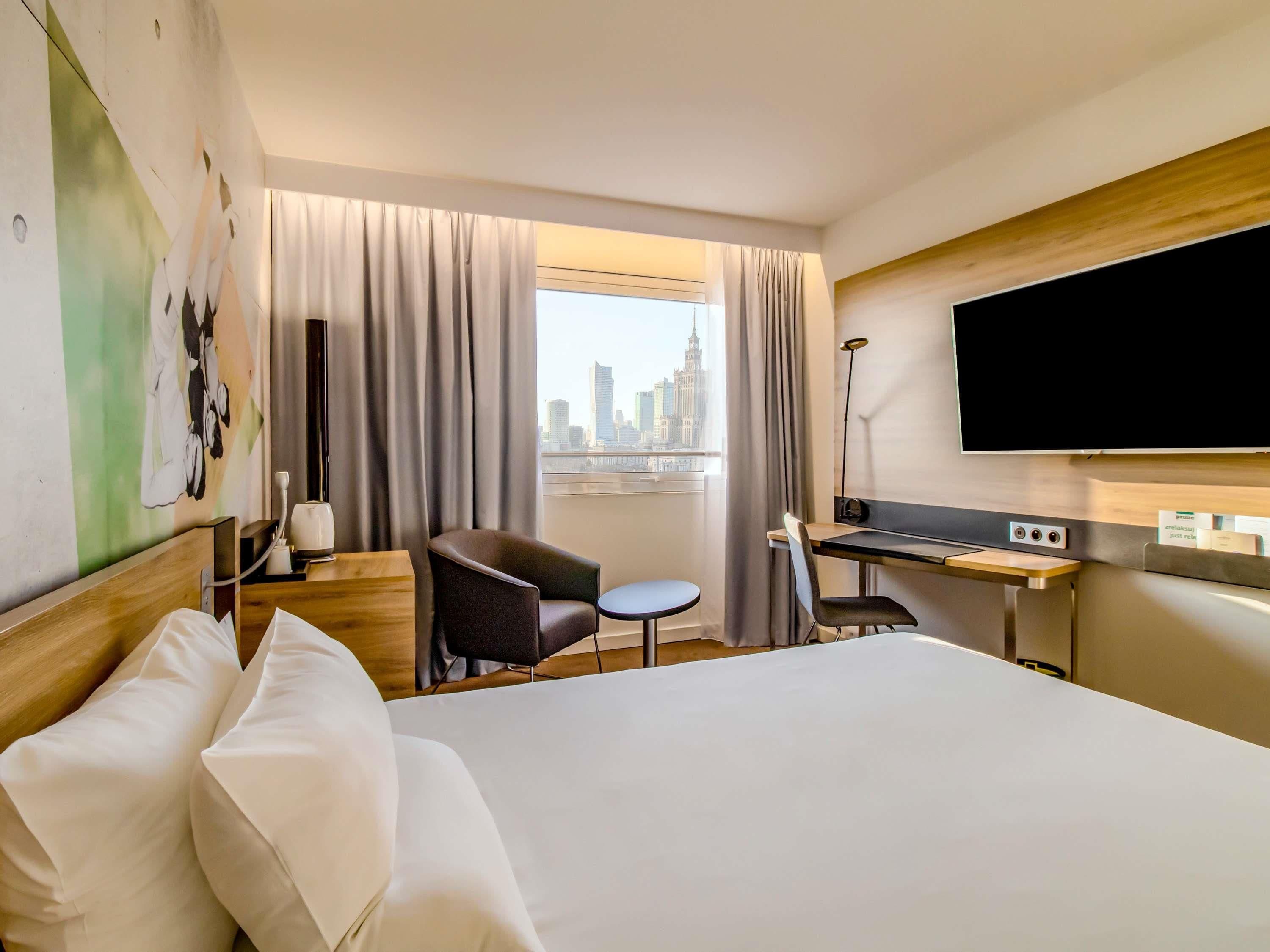 Hotellihuone, jossa on sänky, työpöytä, tuoli, televisio ja näkymä kaupunkimaisemaan.