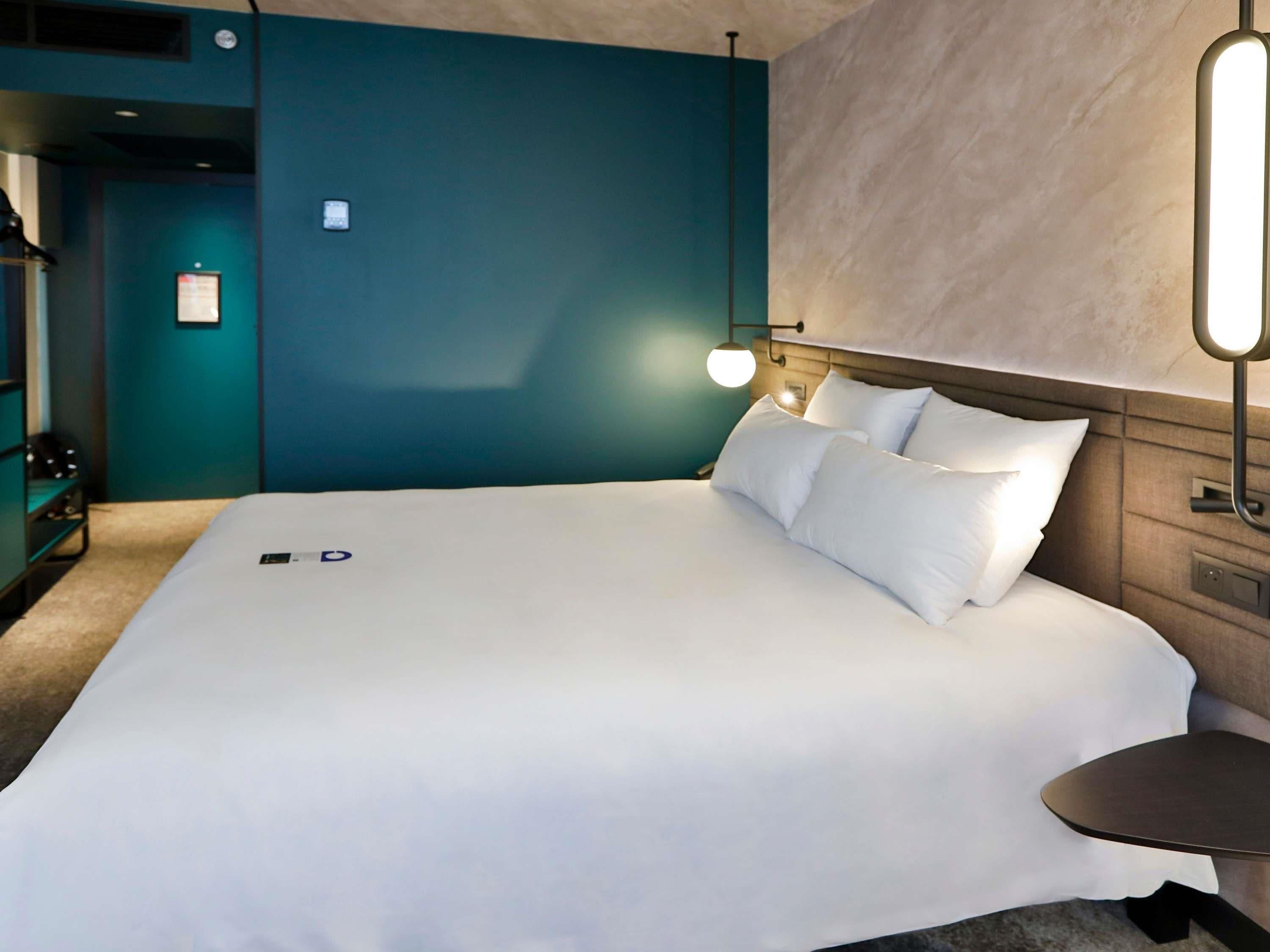 Una habitación de hotel moderna con una cama grande, una mesita de noche, una lámpara de pared y una puerta con un cartel.
