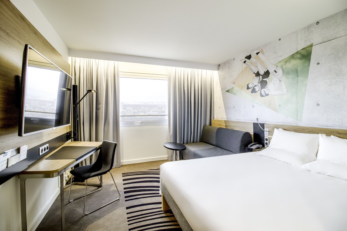 A hotel room with a bed, a desk, a chair, a TV, a phone, and a window with curtains.