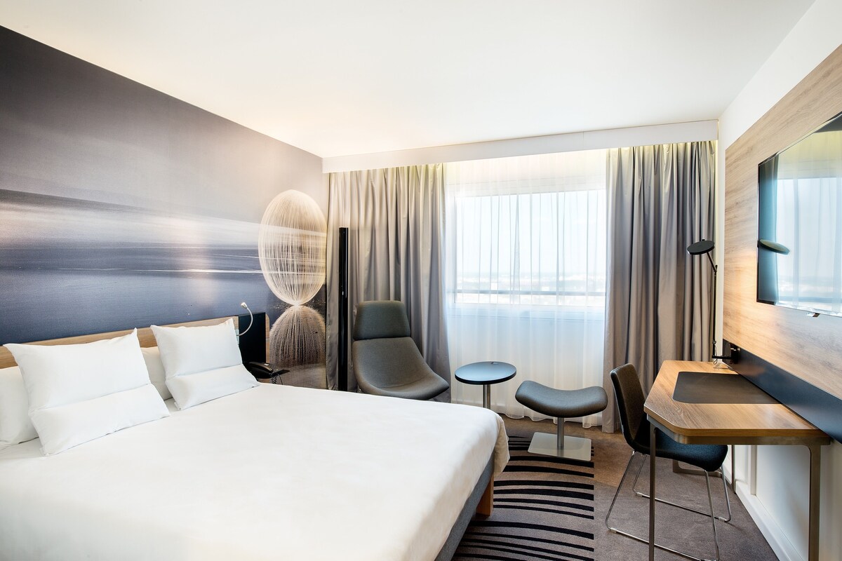 A modern hotel room with a large bed, a desk, a chair, and a small table.