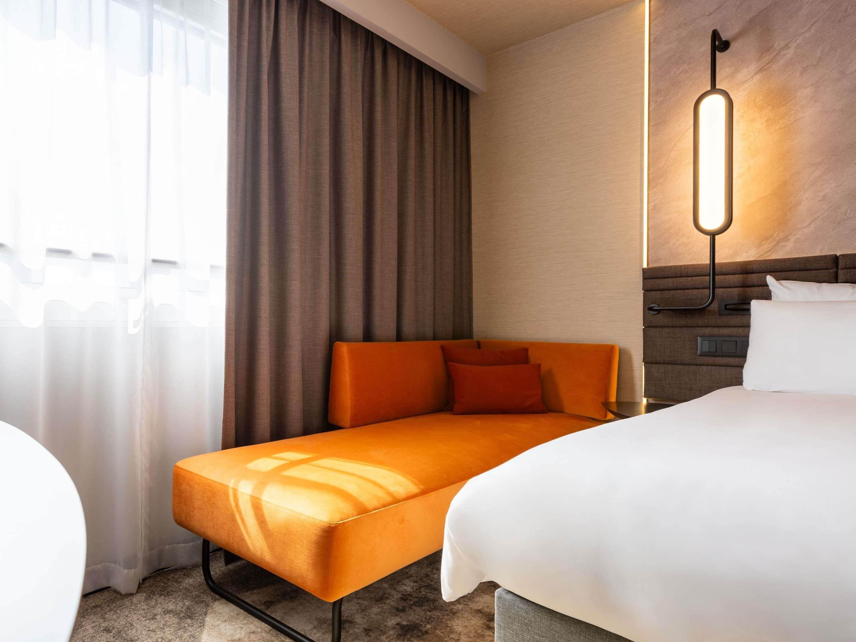 Une chambre d’hôtel avec un canapé-lit, un lit séparé, une lampe fixée au mur et un rideau.