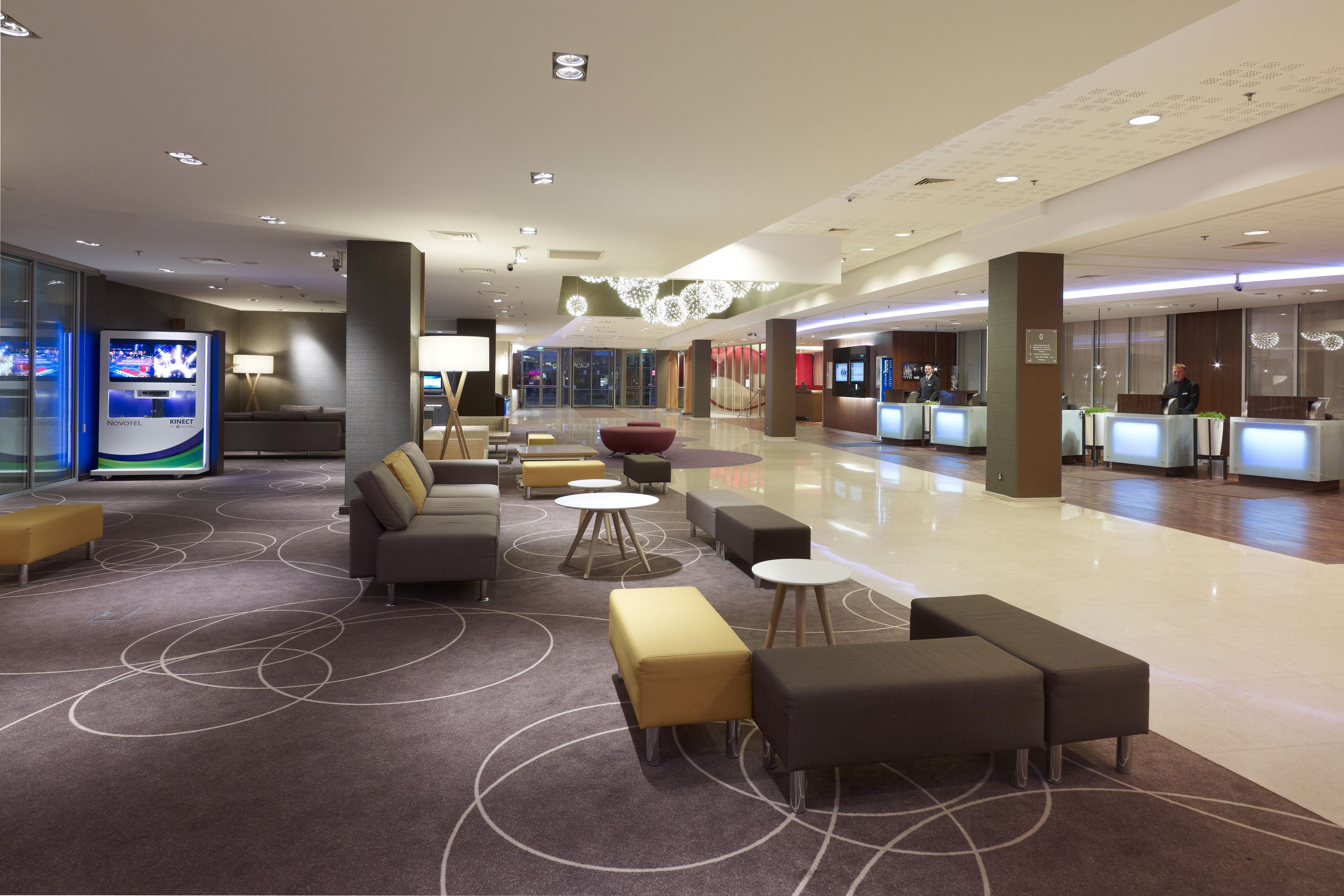 A modern hotel lobby with seating areas, a reception desk, and a digital kiosk.