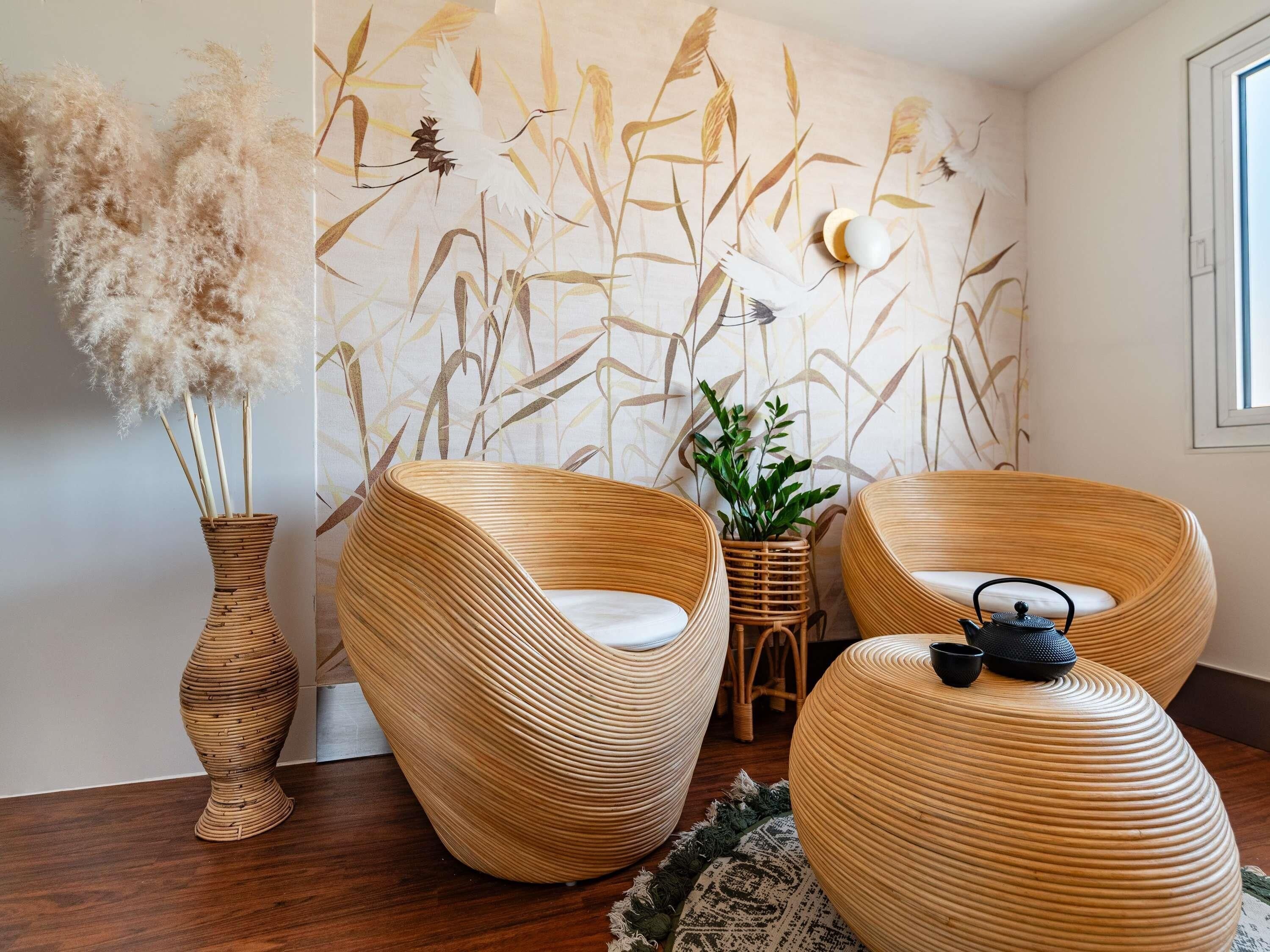 Una habitación con una silla tejida, un taburete tejido, un jarrón con plumas de mispira y una pared con papel tapiz de motivos naturales.
