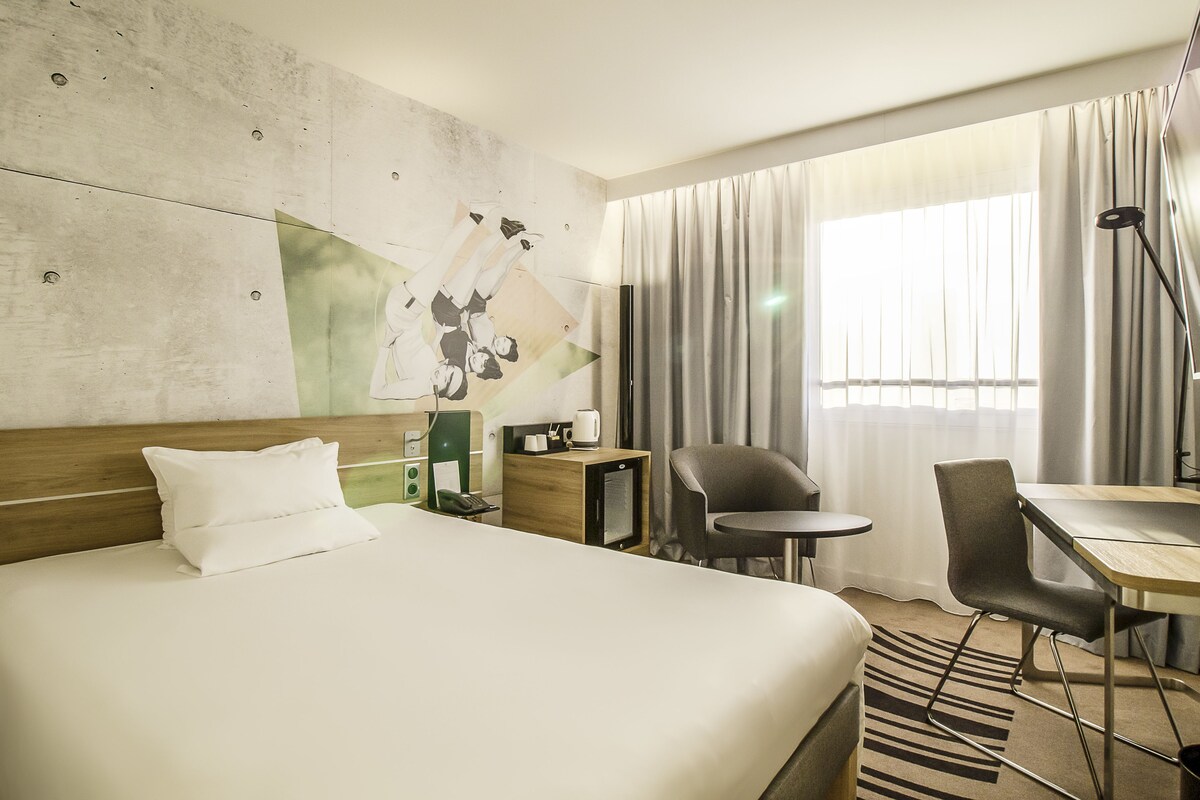 A hotel room with a large bed, a desk, a chair, a small table, and a wall-mounted TV.