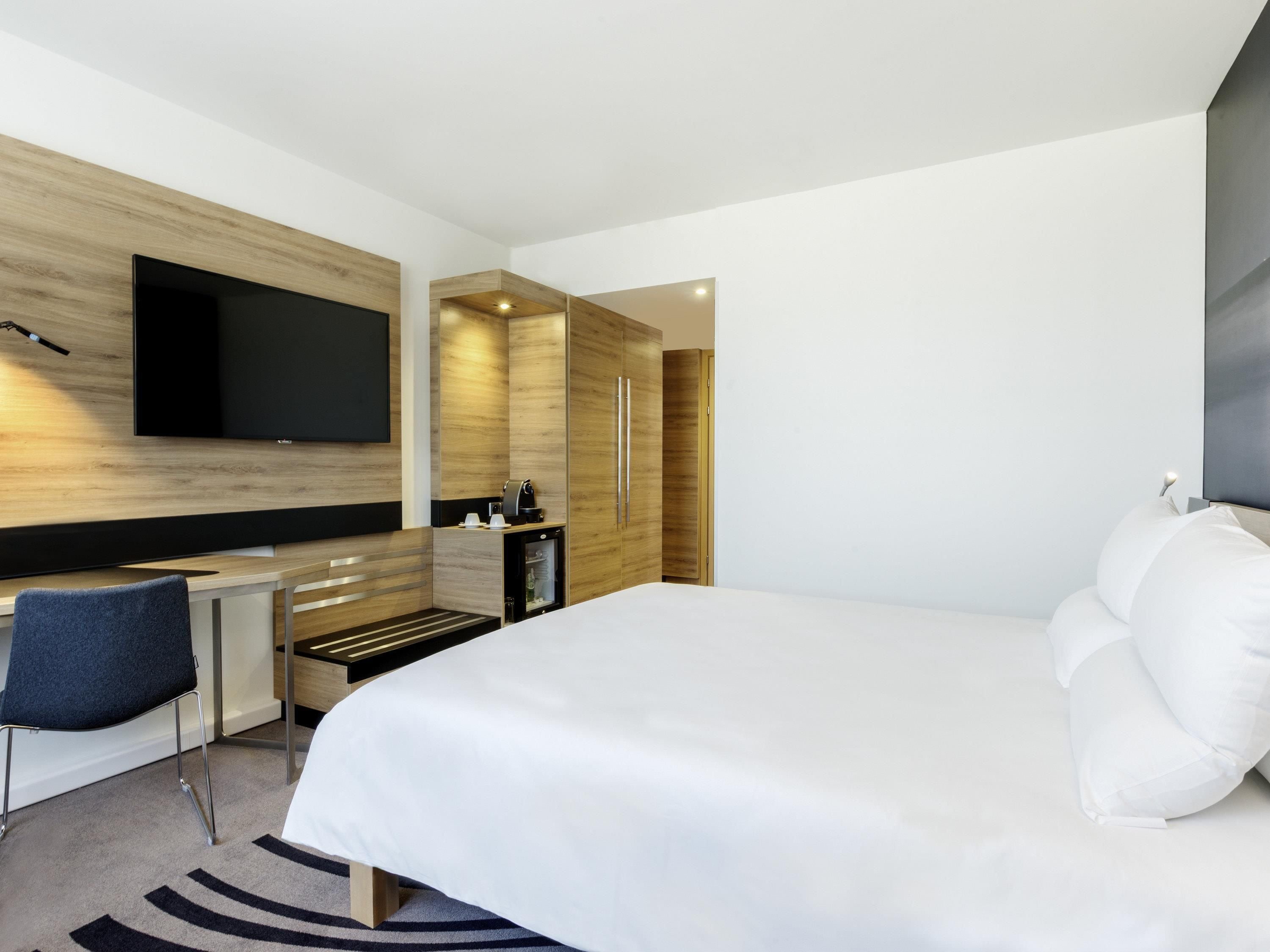 Een moderne hotelkamer met een groot bed, een flatscreen televisie aan de muur, een kleine kitchenette en een zithoek met een stoel.