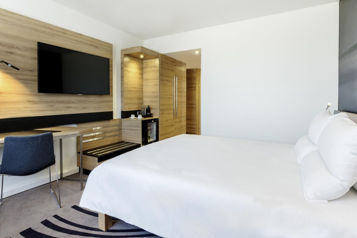 A modern hotel room with a large bed, a flat-screen TV mounted on the wall, a small kitchenette, and a seating area with a chair.