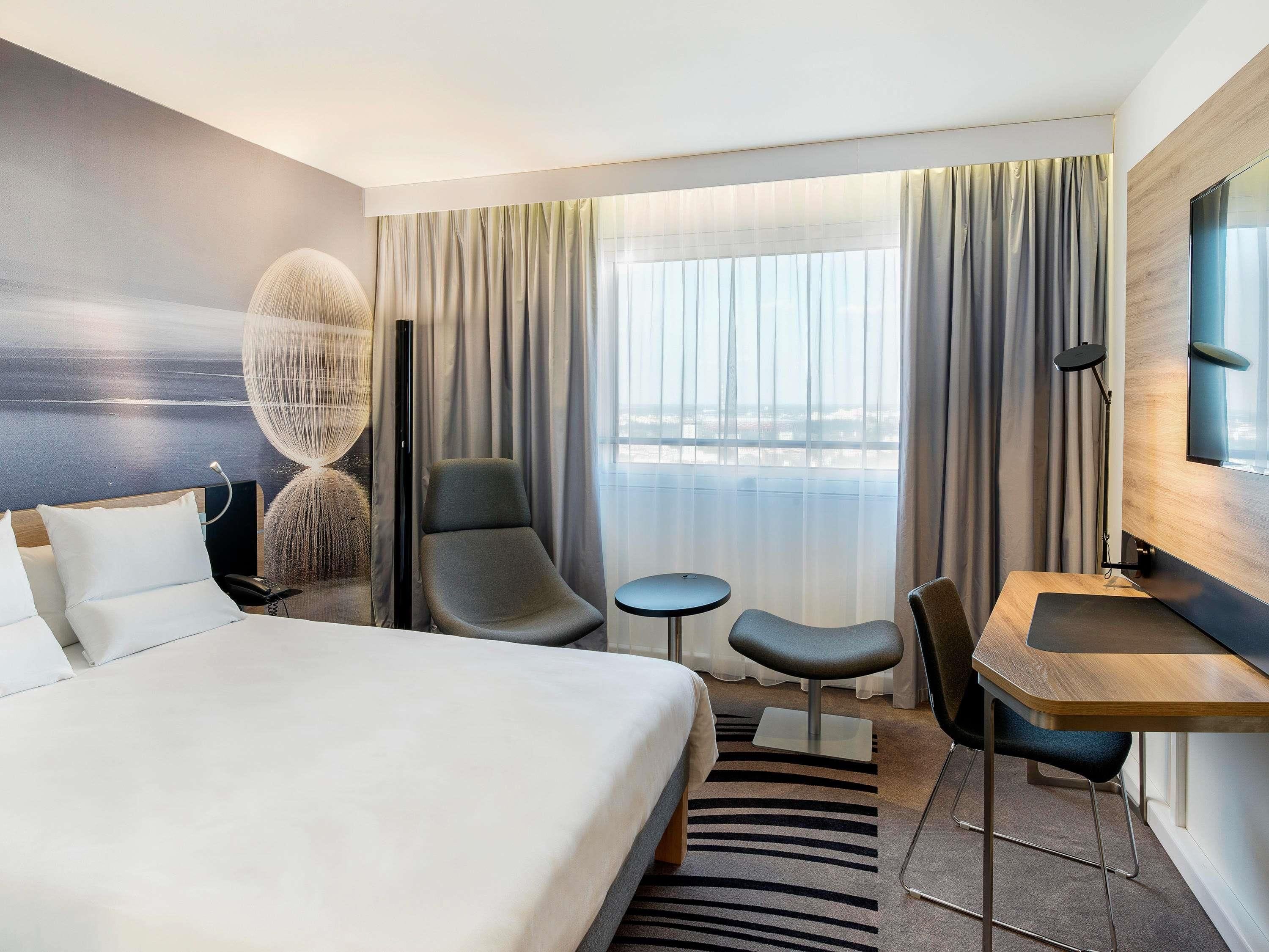 Habitación de hotel con cama, escritorio, silla, mesita pequeña y un ventanal grande con cortinas.