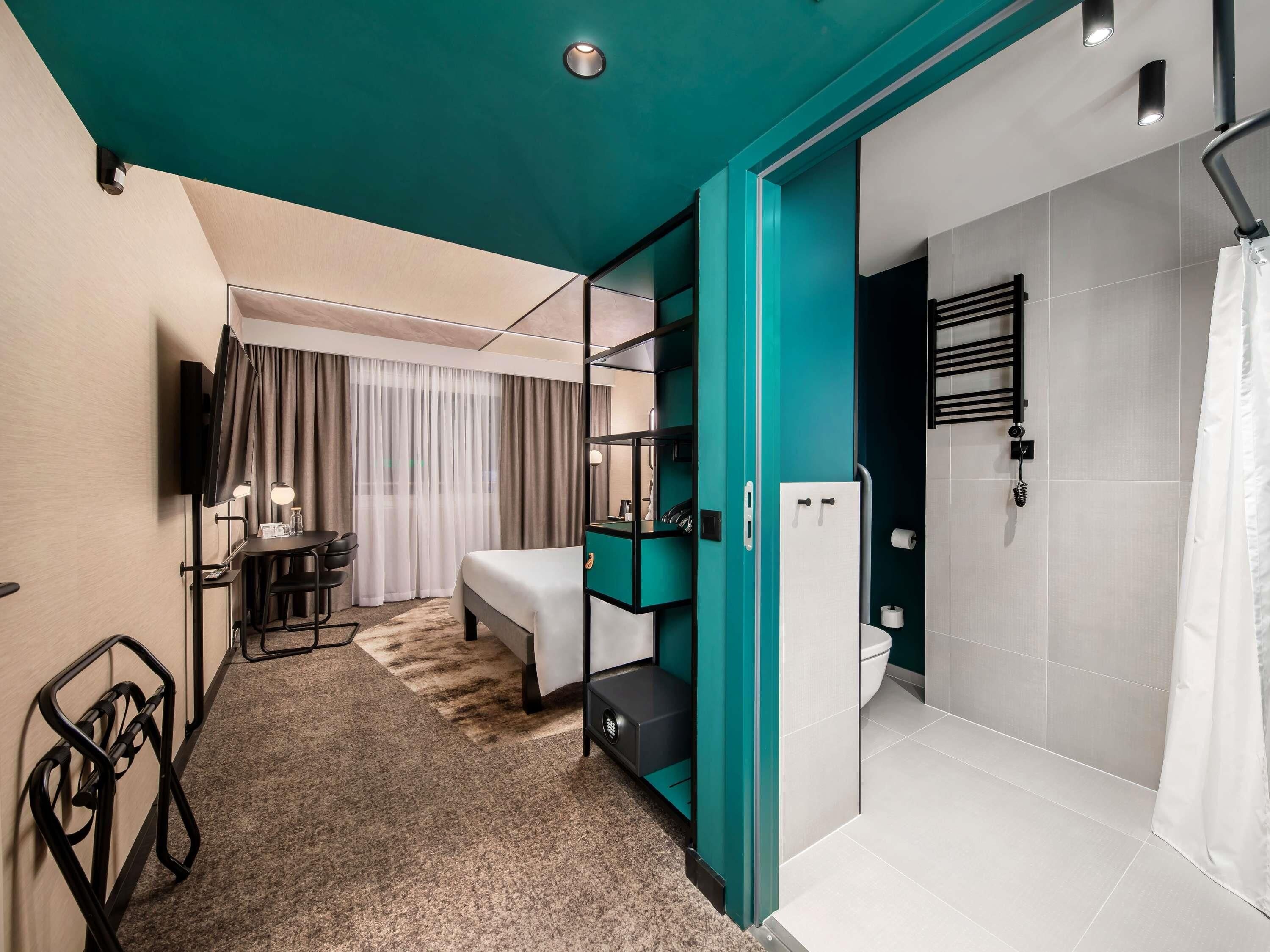 A modern hotel room with a bed, a desk, a TV, a shower, and a bathroom with a towel rack.